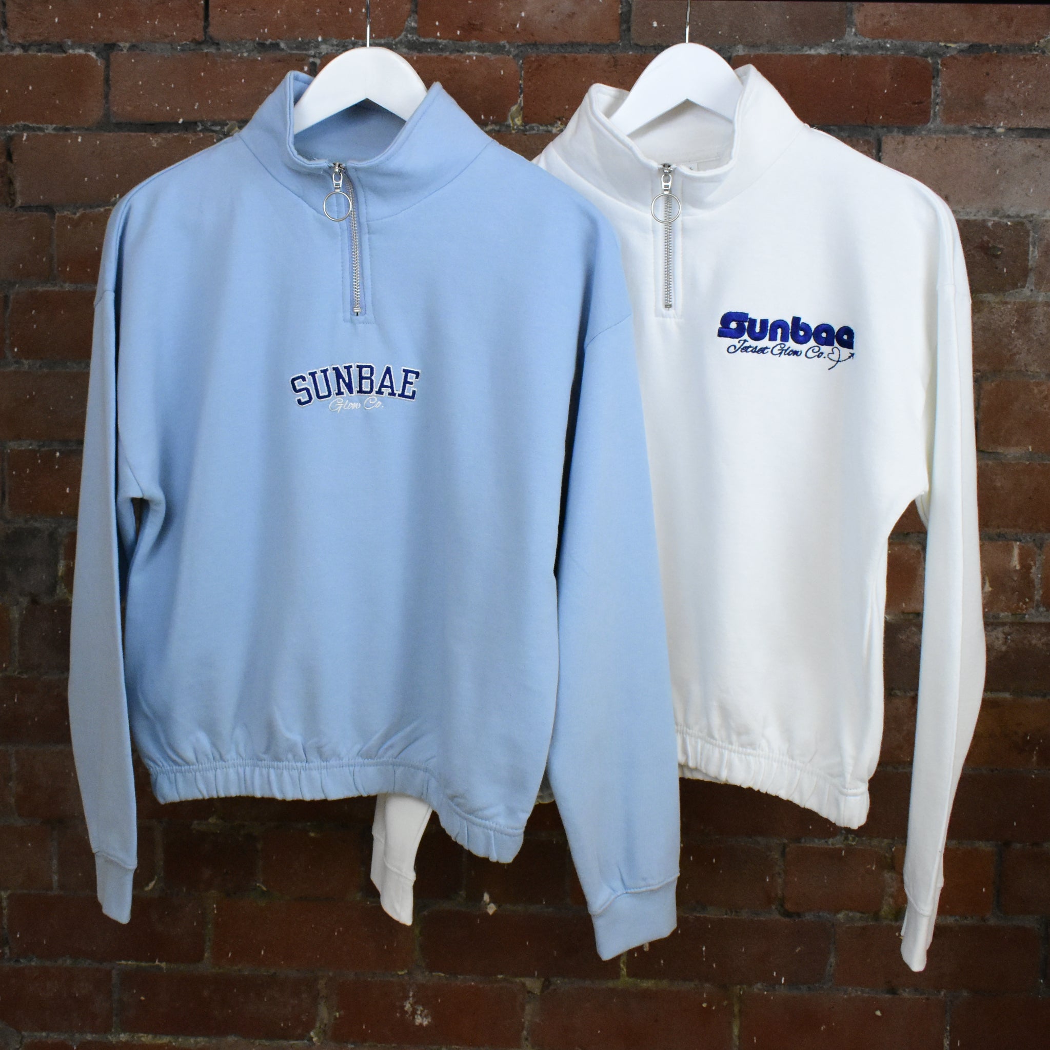 Two sweatshirts, one light blue and one white, with 'SUNBAE' branding on hangers against a brick wall.