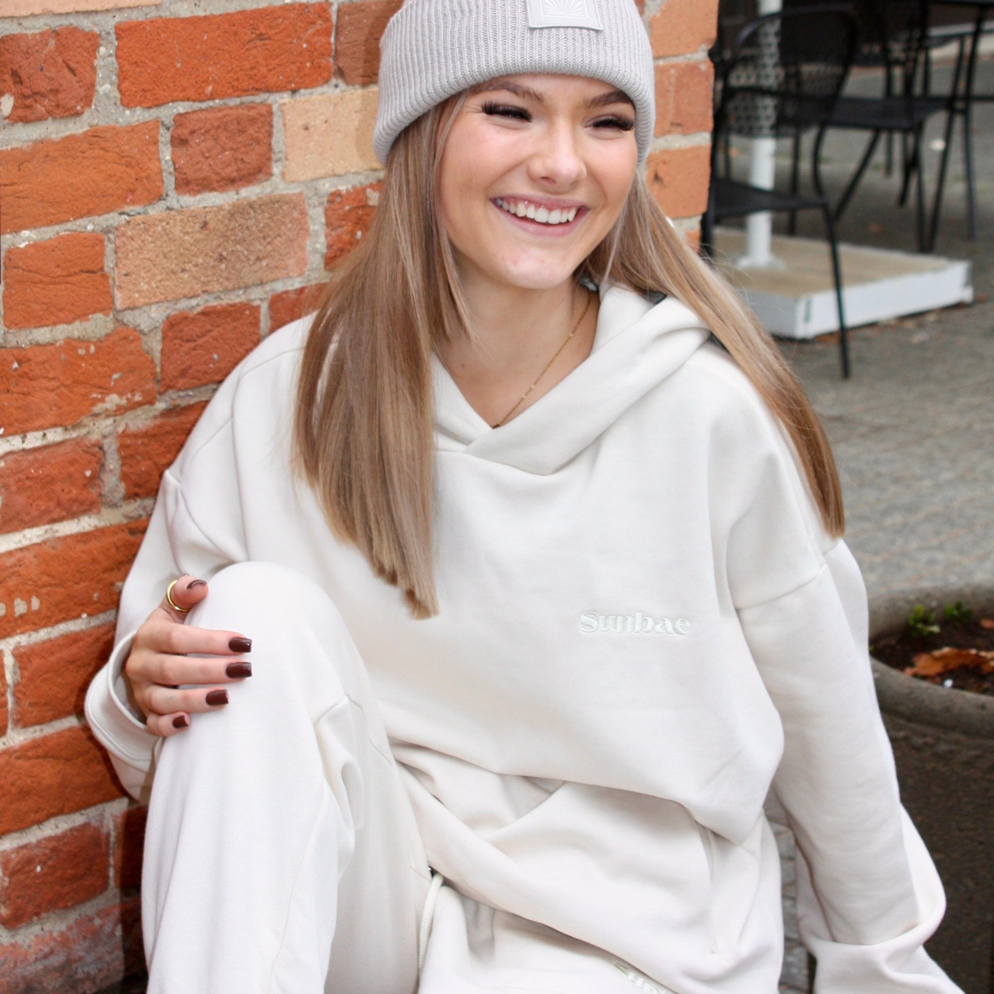 Person wearing a white hoodie and gray beanie sitting against a brick wall.