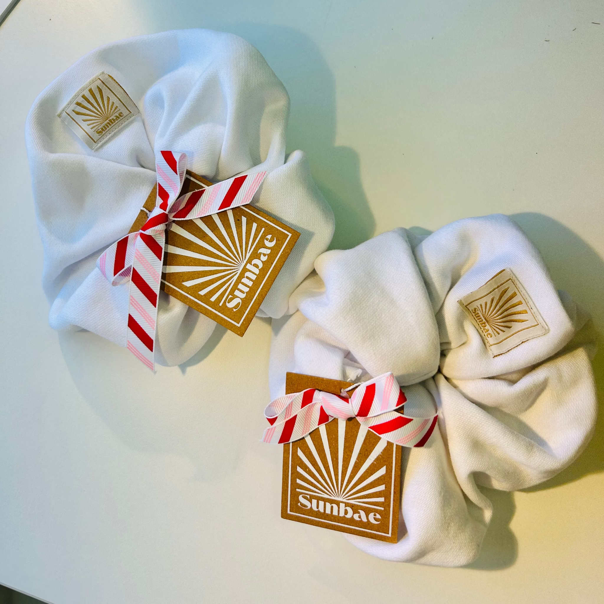 White SUNBAE hair scrunchies with candy cane ribbons and SUNBAE tags on a light surface.