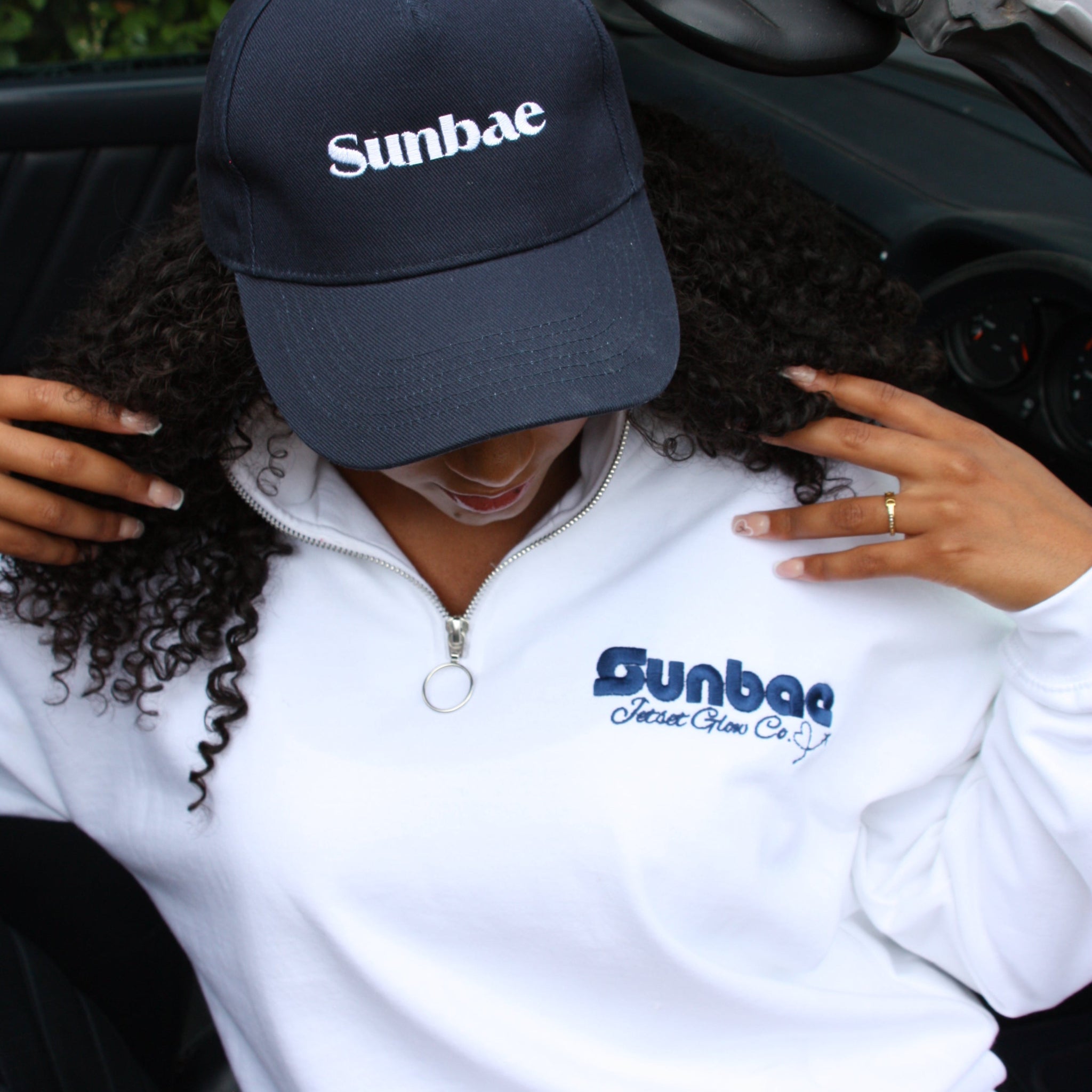 Person wearing a white hoodie and navy cap with 'Sunbae' branding, leaning over a vehicle.