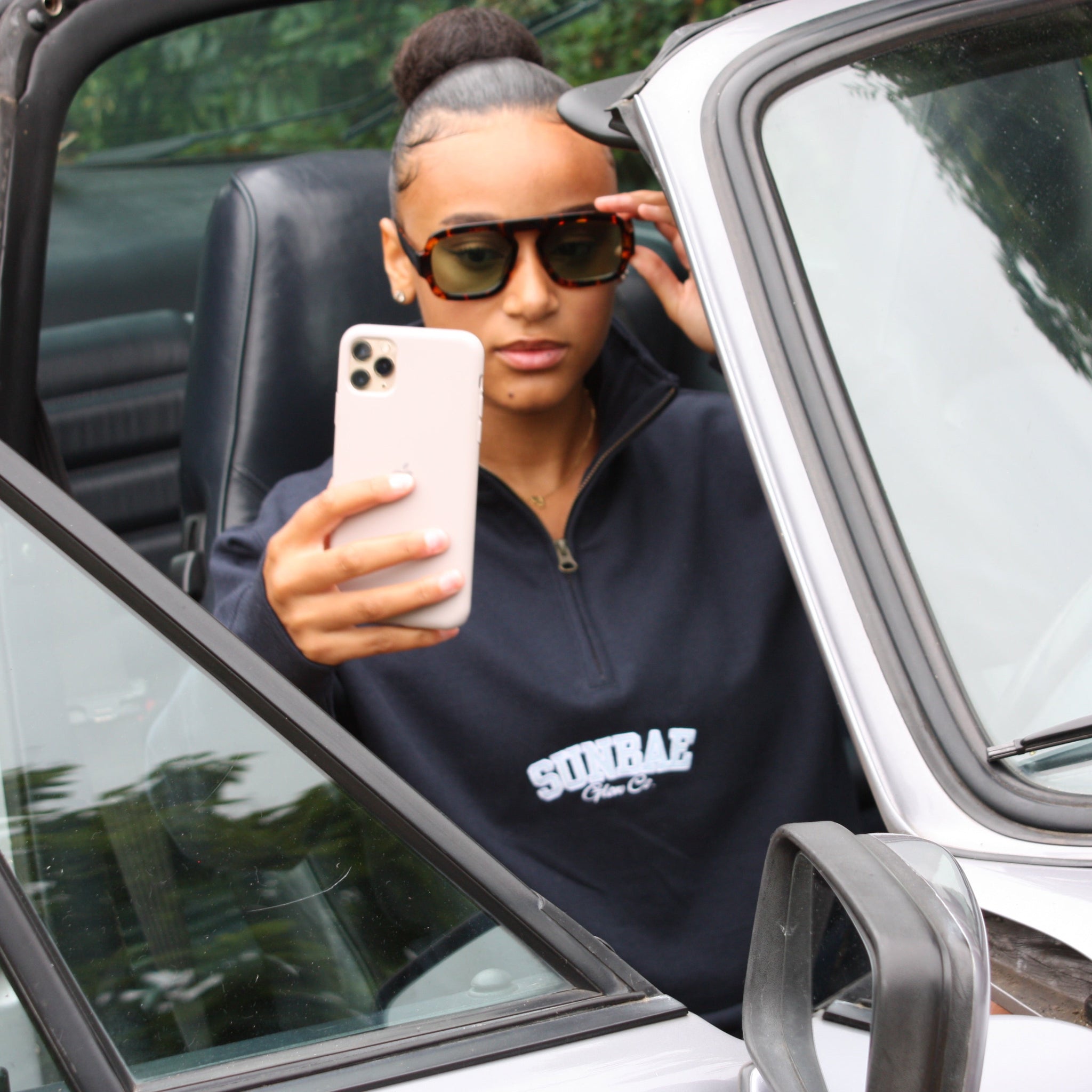 Person taking a selfie in a car wearing sunglasses and a dark hoodie with 'SUNBAE' text.