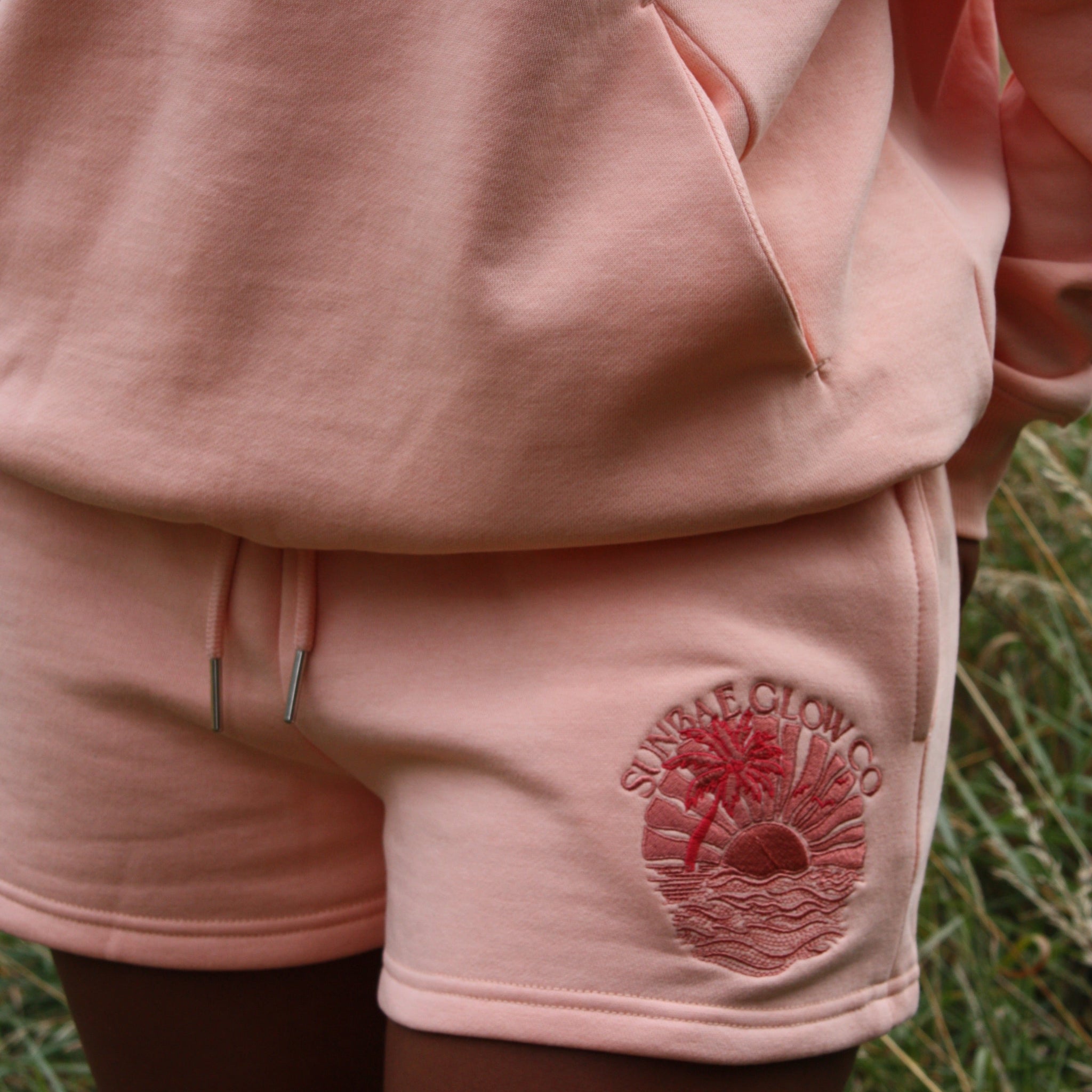 Person wearing a coral Sunbae Co-Ord with a logo on shorts against a grassy background