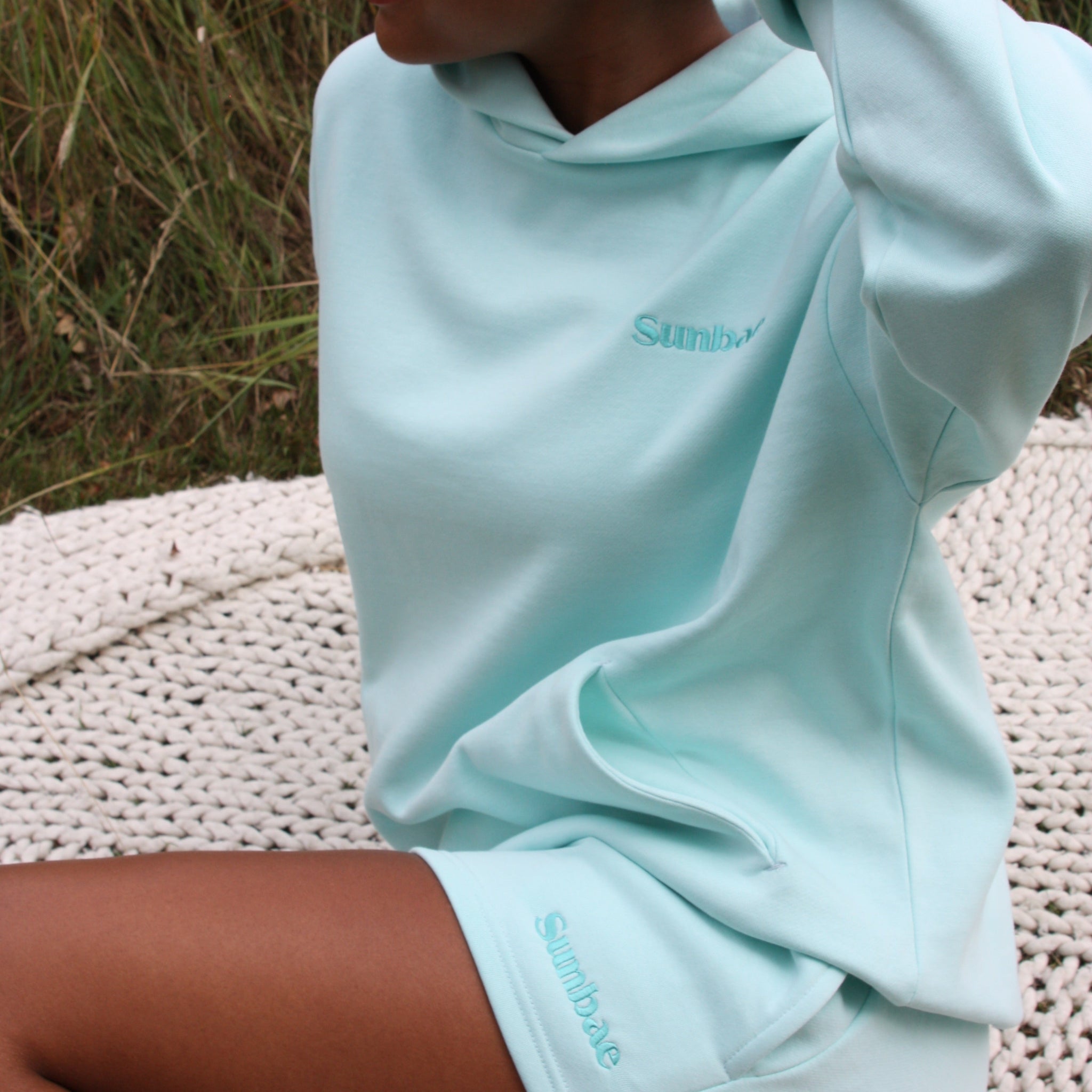 Person wearing mint Sunbae hoodie sitting on a textured surface with grass in the background.
