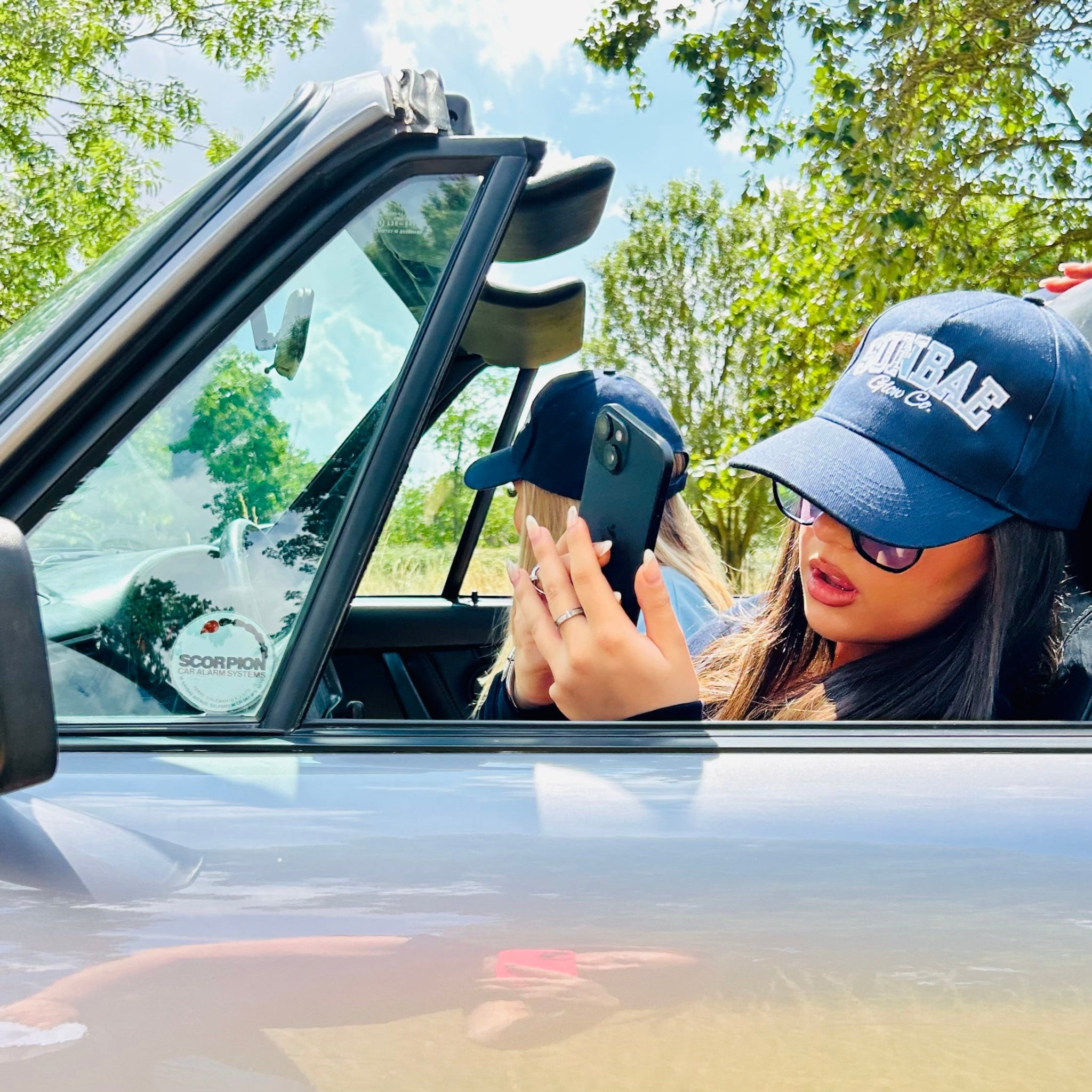 Sunbae Glower in a car wearing Sunbae Solar Cap and sunglasses, looking at a phone.