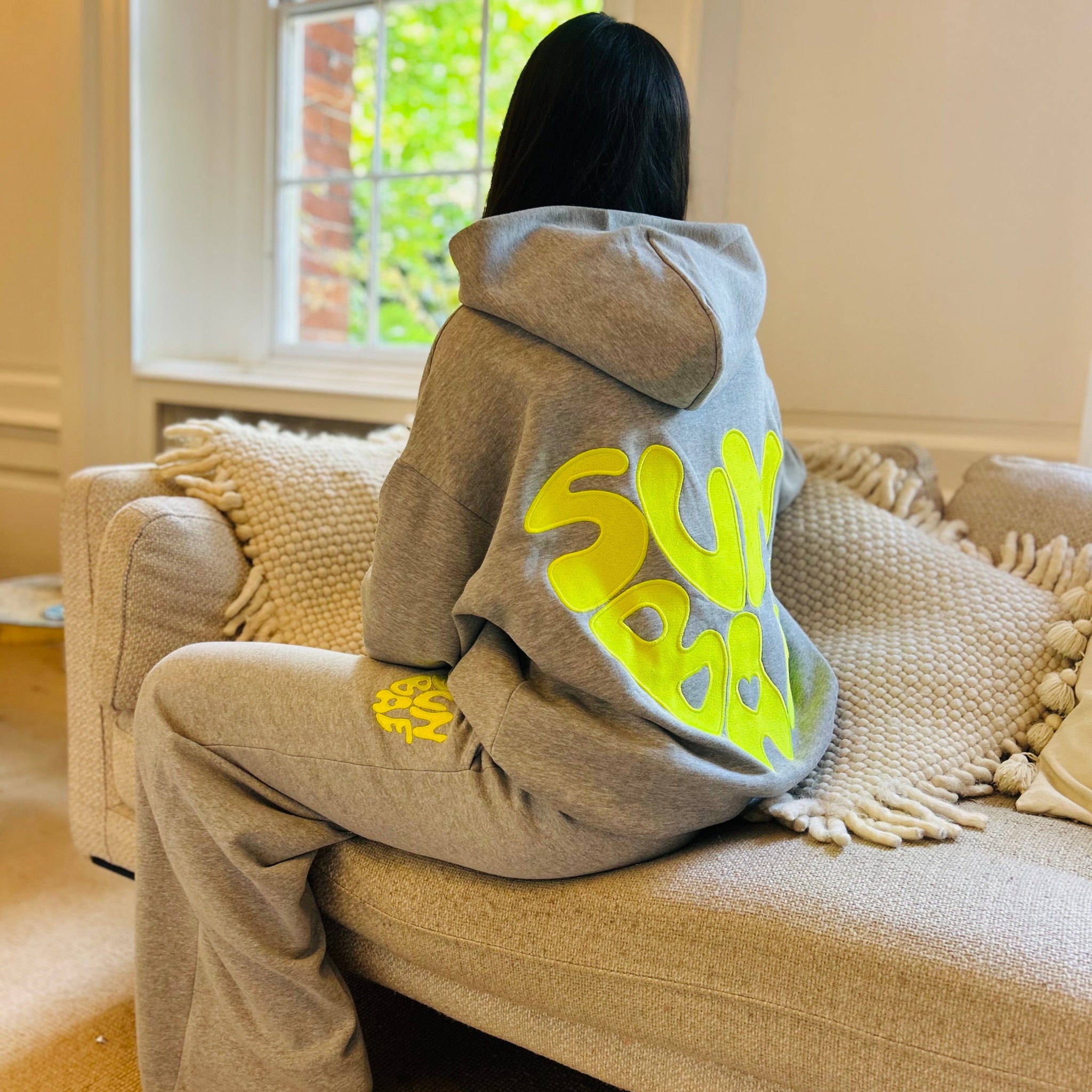 Sunbae Glower wearing SUNBAE Offline Oversize Hoodie with Glazed Heart Embroidery in Grey Marl sitting on a beige couch.