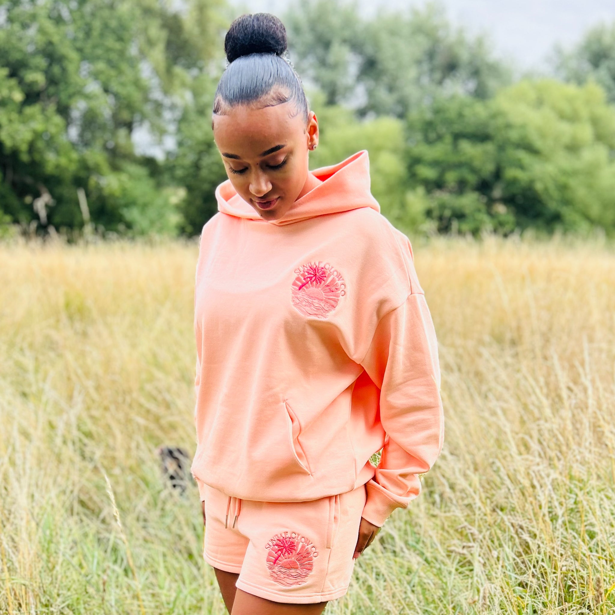 Person wearing a coral Sunbae hoodie and shorts set with a pink floral design in a grassy field.