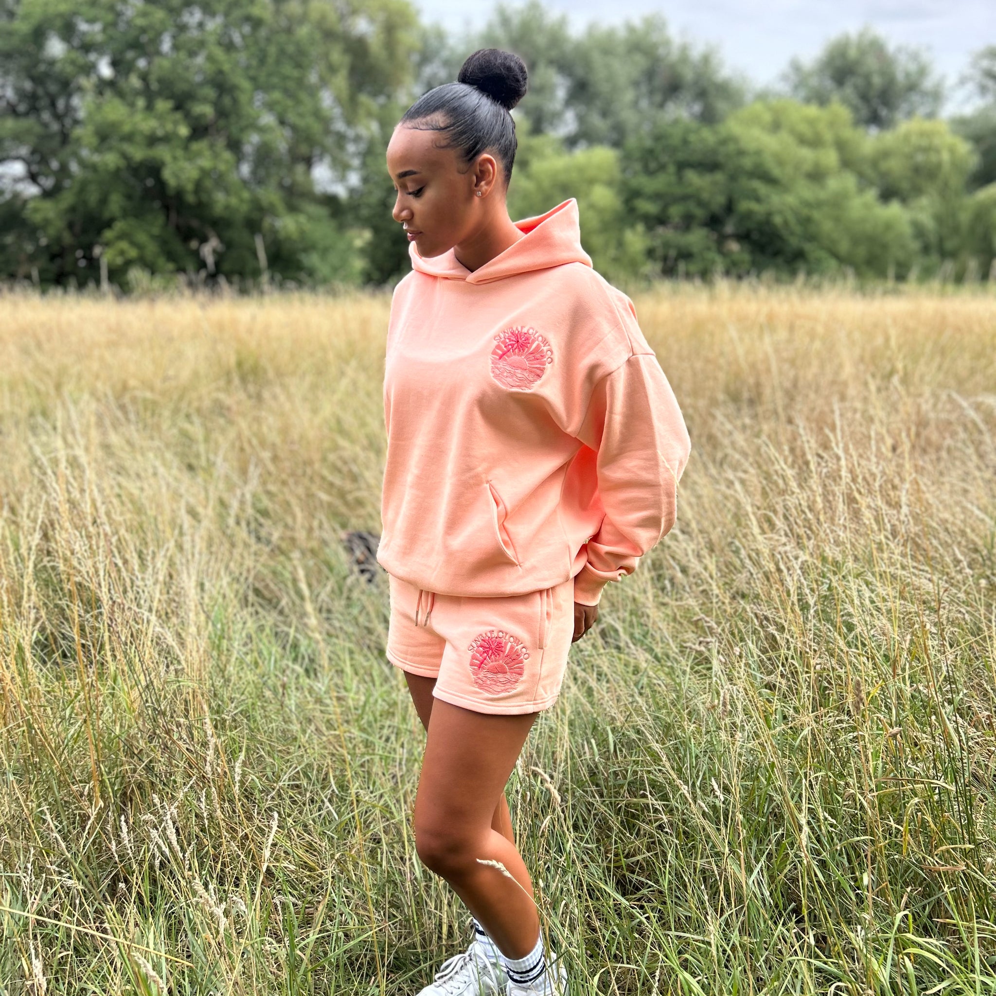 Person wearing a coral Sunbae hoodie and shorts set in a field with trees in the background