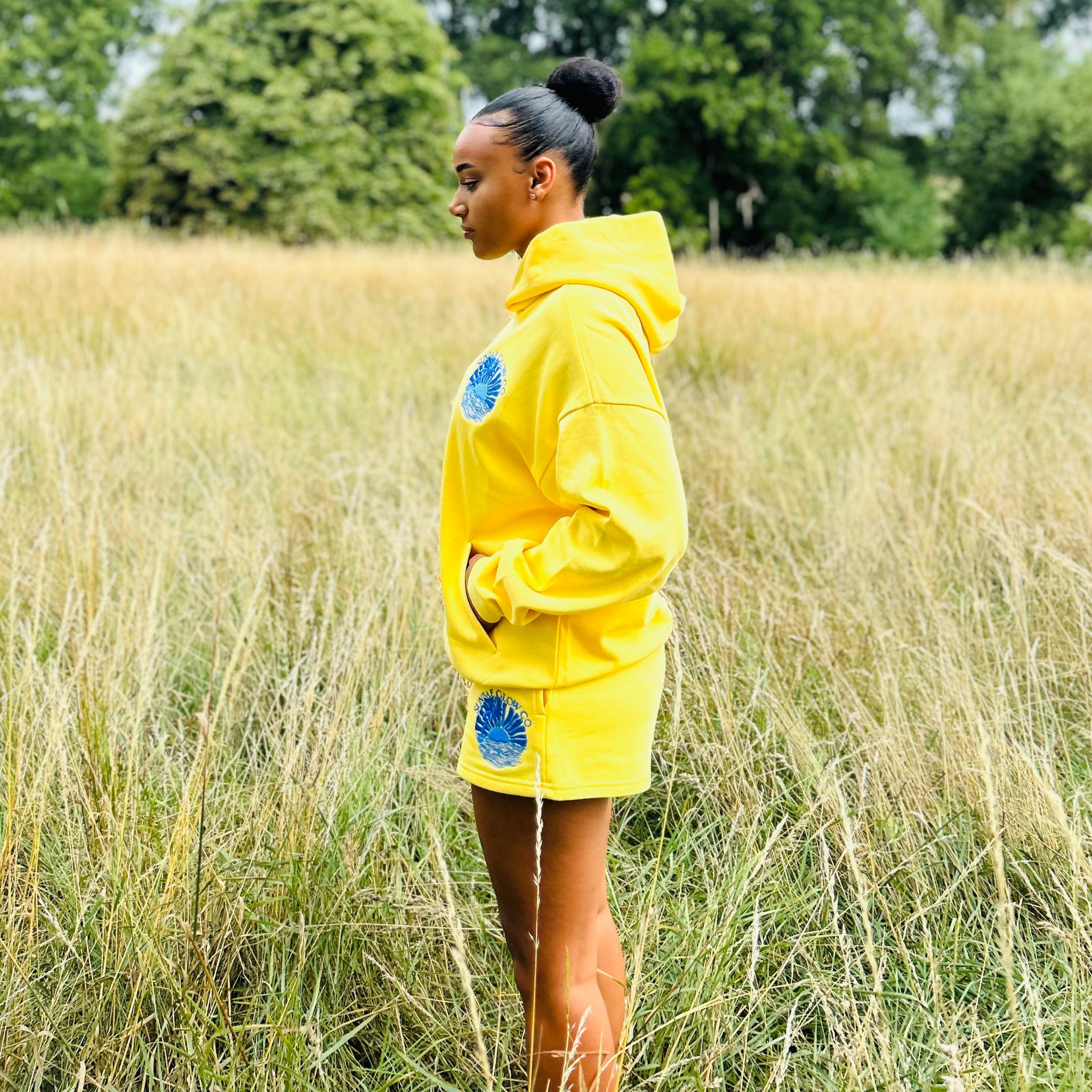 Person wearing a yellow Sunbae hoodie with blue designs standing in a grassy field.