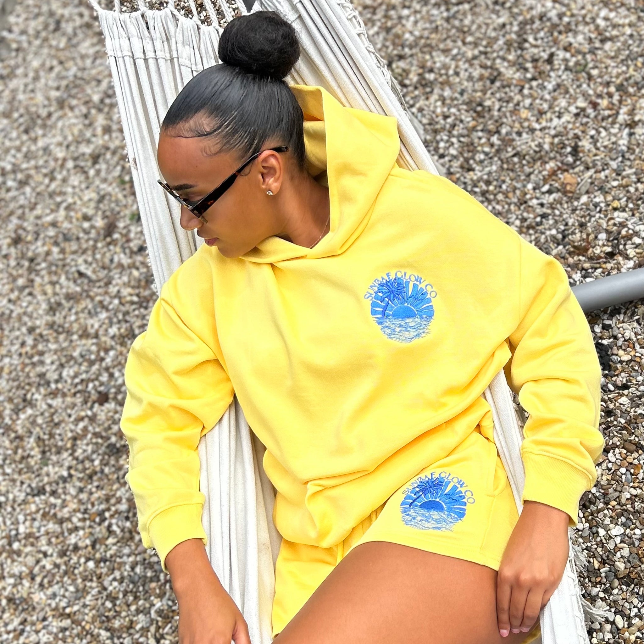 Person wearing a yellow Sunbae hoodie with a blue logo, sitting on a hammock.