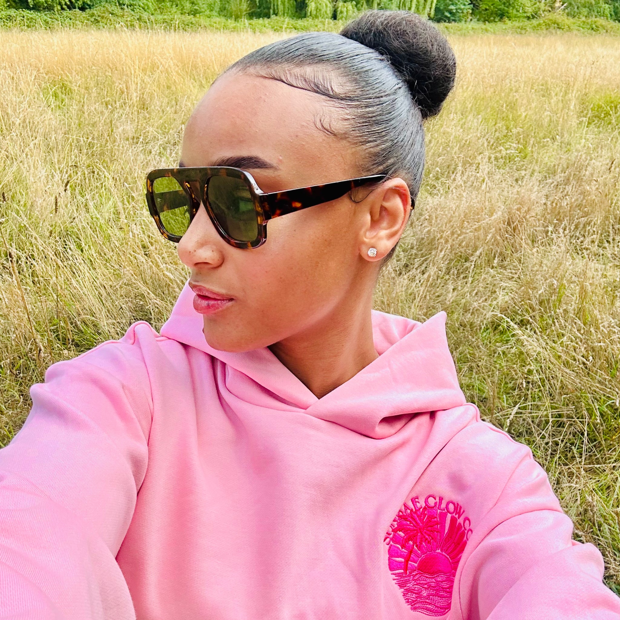 Person wearing a pink Sunbae hoodie with sunglasses and a bun in a grassy field