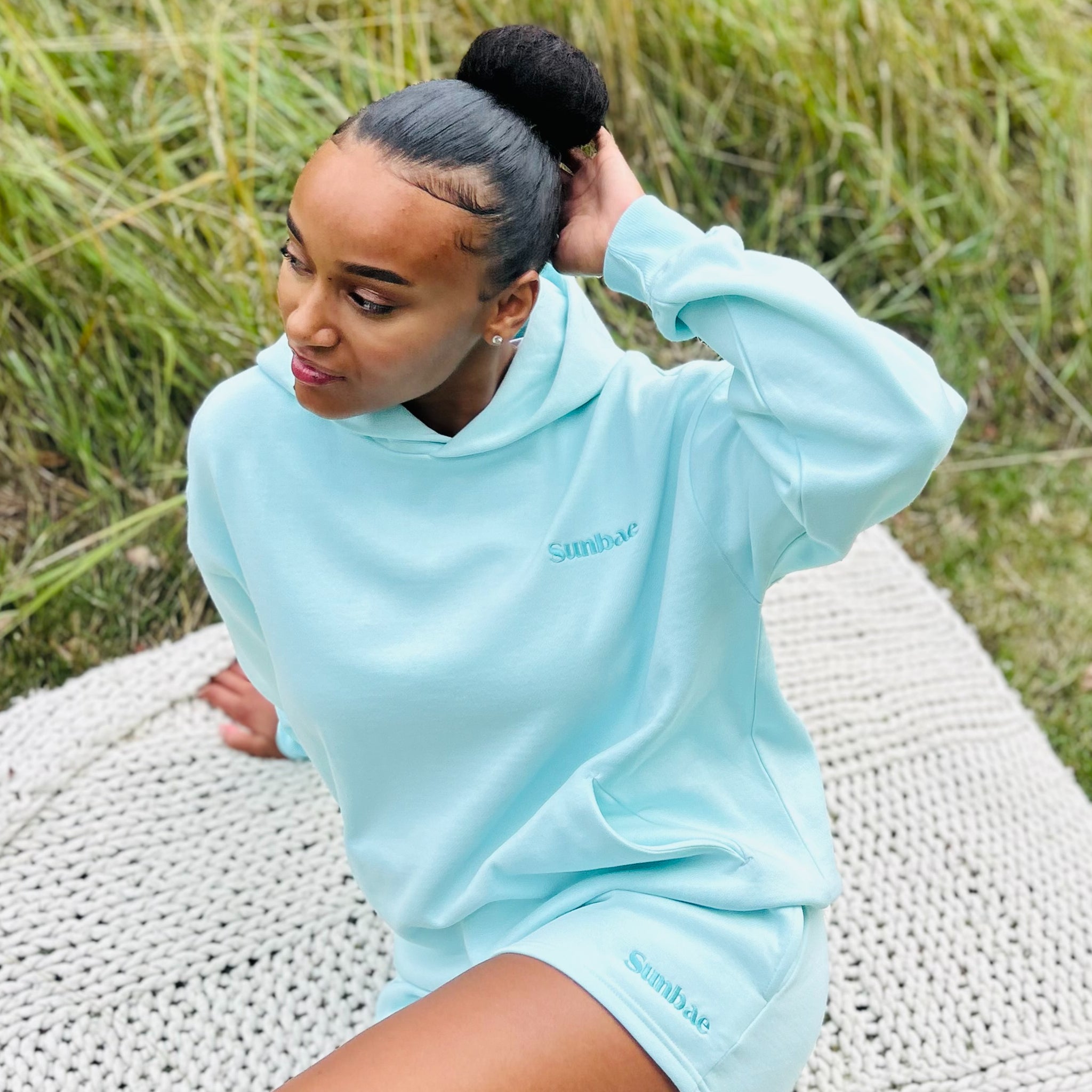 Woman wearing a mint Sunbae hoodie and short sitting on a woven mat outdoors.