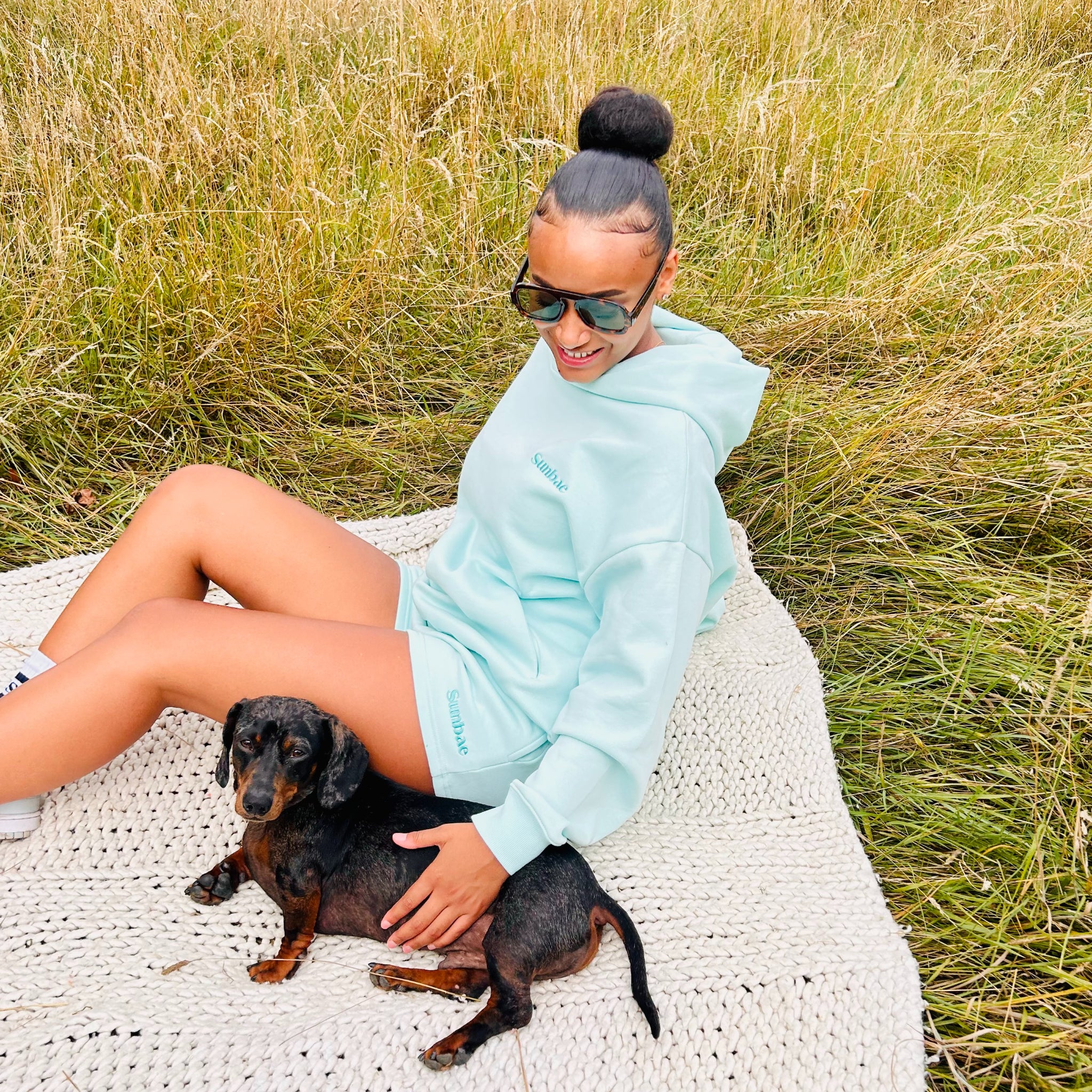 Woman in mint Sunbae hoodie sitting on a blanket with a small dog in a grassy field