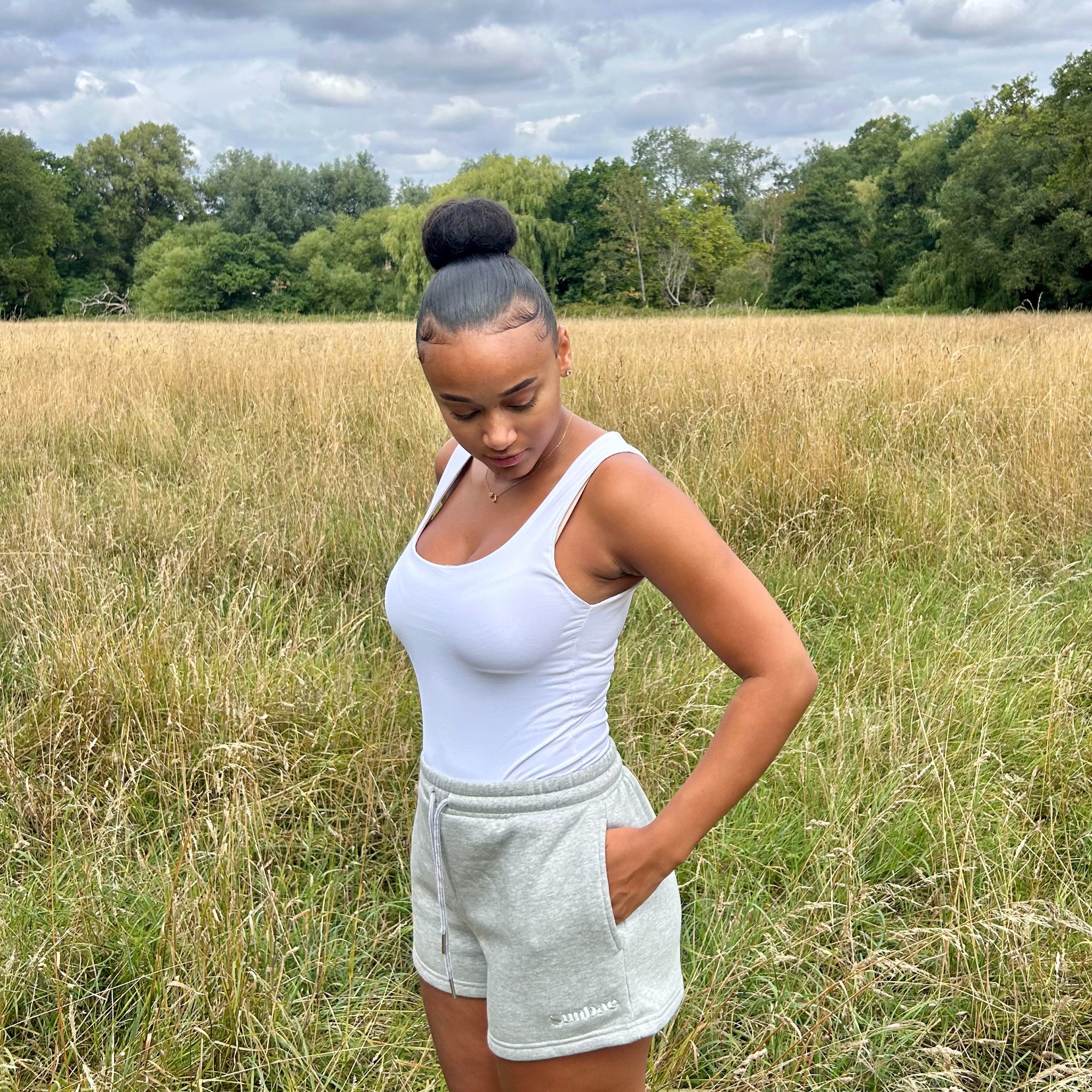 Woman in a white tank top and grey Sunbae shorts standing in a field with trees in the background