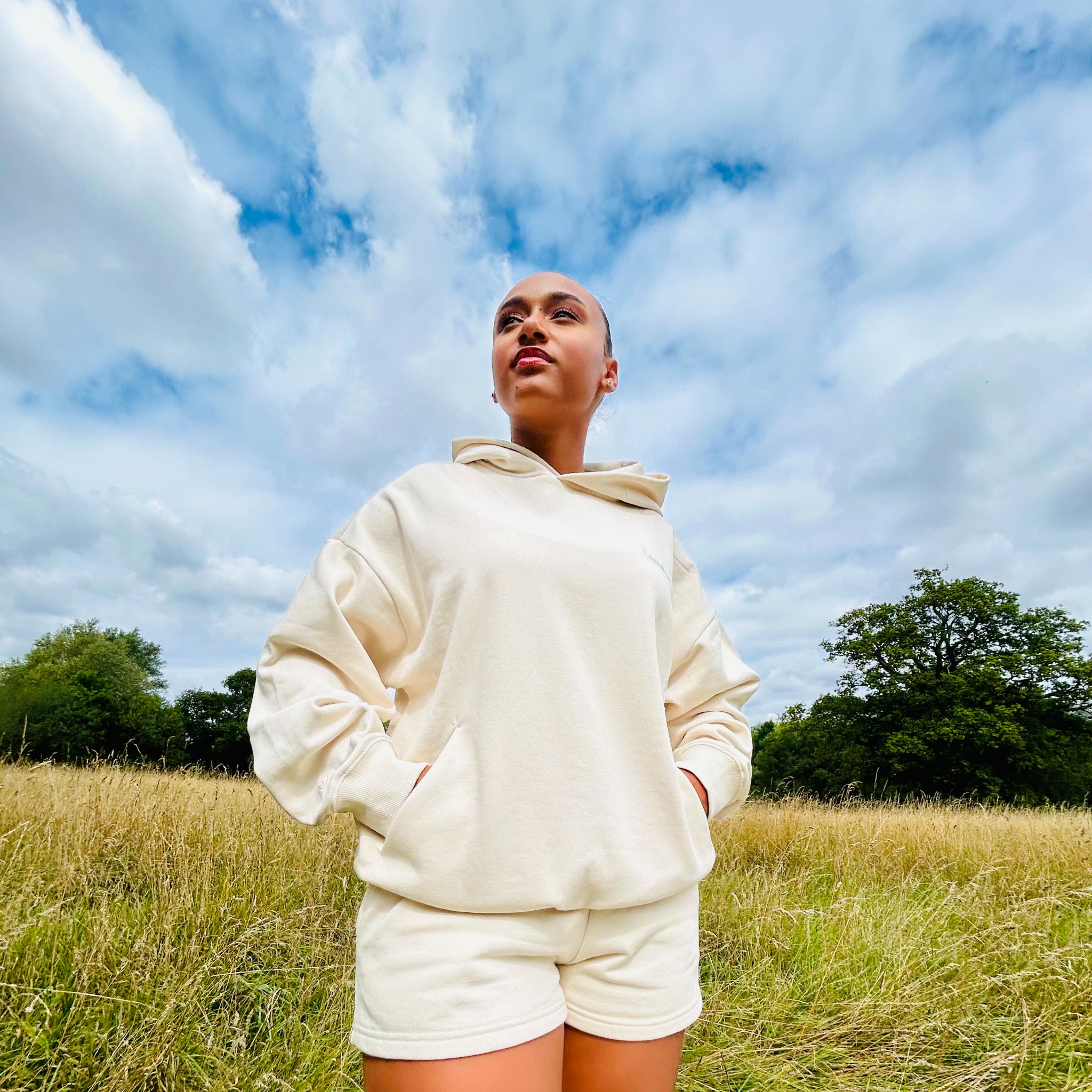 Person wearing a ivory Sunbae hoodie and shorts standing in a field with a blue sky.