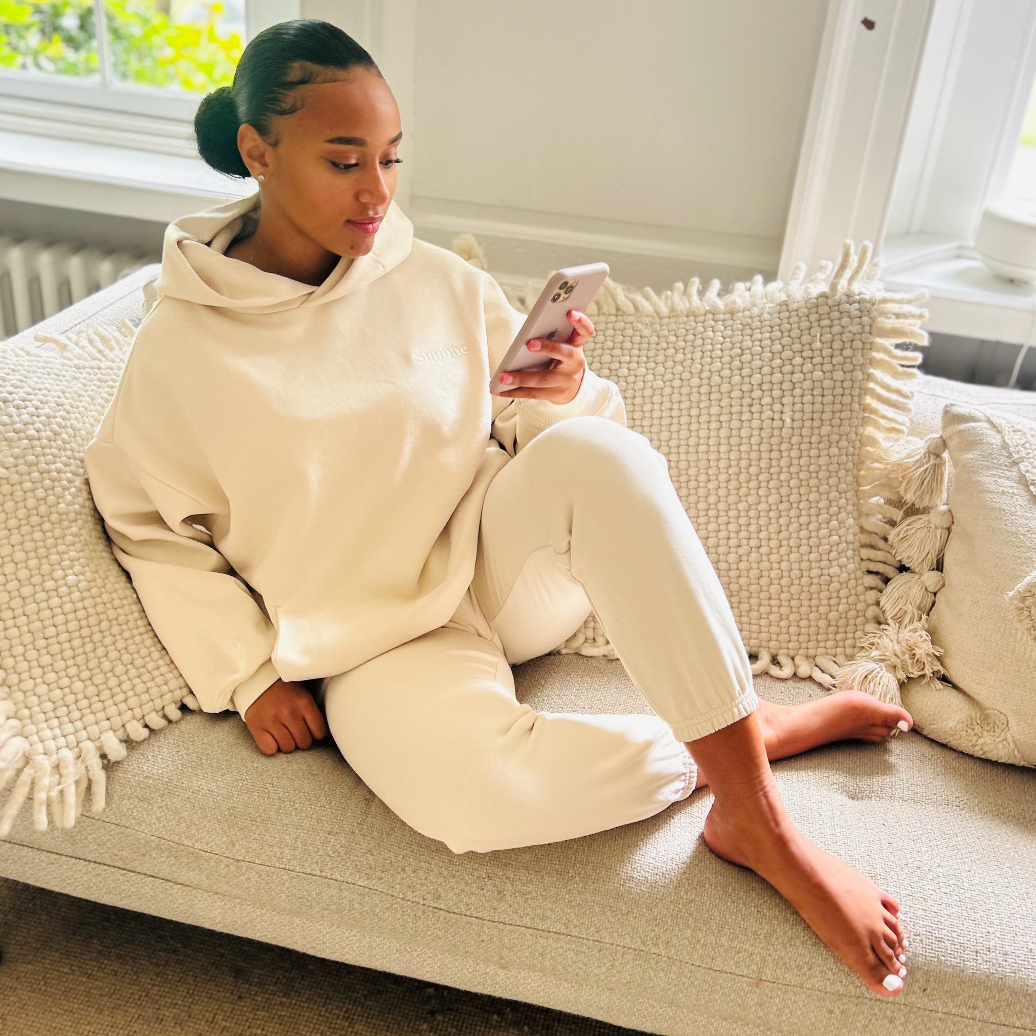 Woman in an ivory Sunbae hoodie and pants sitting on a couch, using a phone.