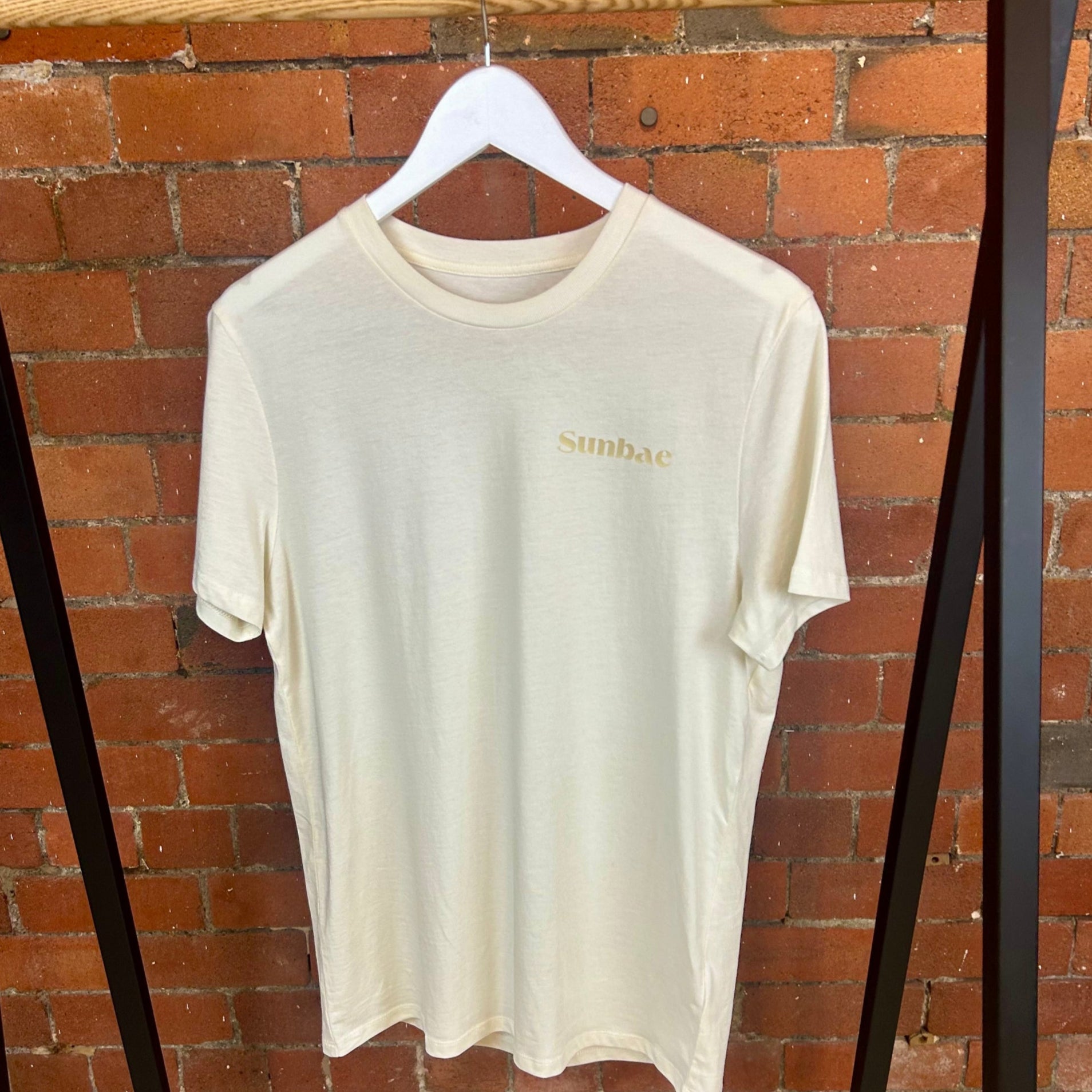 Beige t-shirt with 'SUNBAE' branding on a hanger against a brick wall.