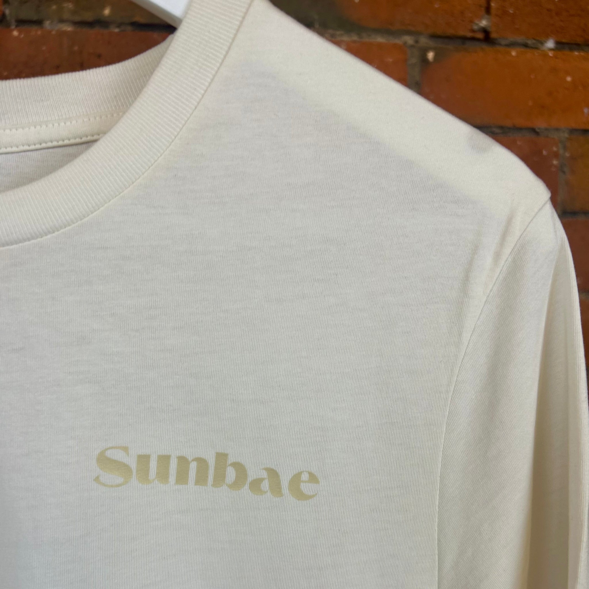 Beige t-shirt with 'Sunbae' text on a hanger against a brick wall.
