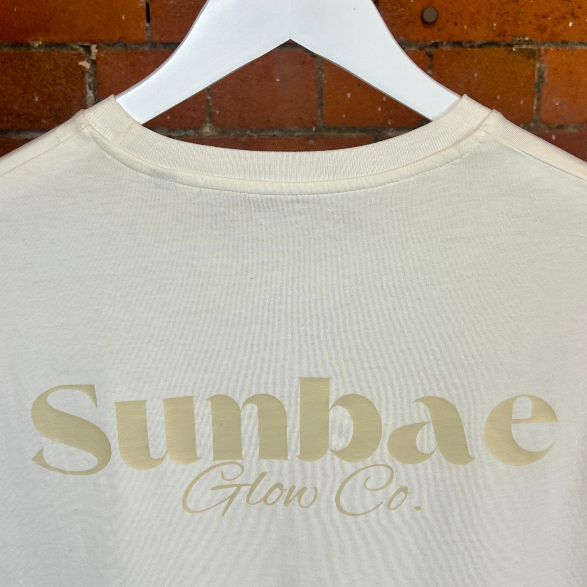 Beige t-shirt with 'Sunbae Glow Co.' text on a white hanger against a brick wall.