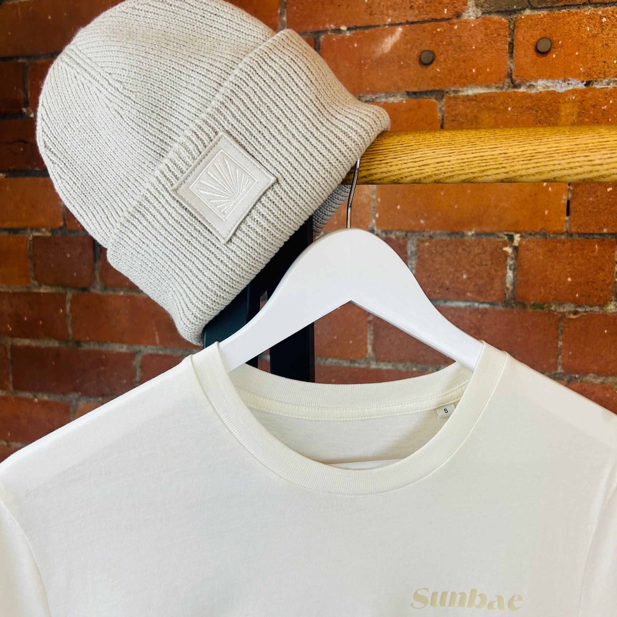Beige knit beanie on a hanger with a white sweatshirt featuring 'Sunbae' branding against a brick wall.