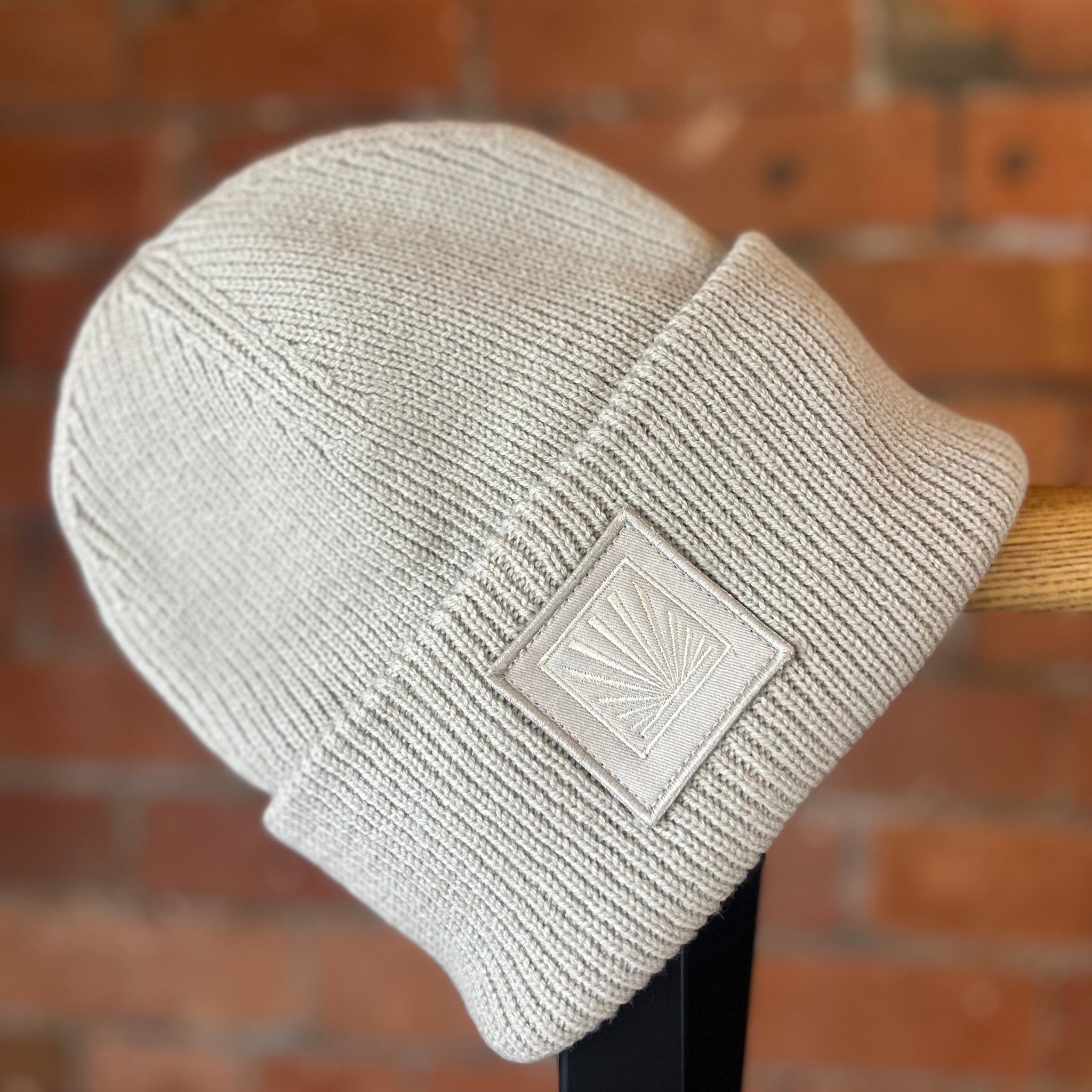 Beige knit SUNBAE beanie with a logo on a wooden stand against a brick wall.