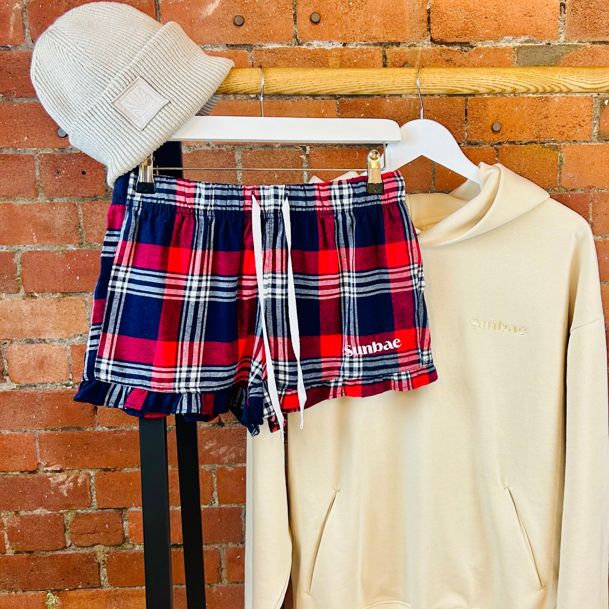 Plaid SUNBAE shorts, beige hoodie, and beanie on a rack against a brick wall.