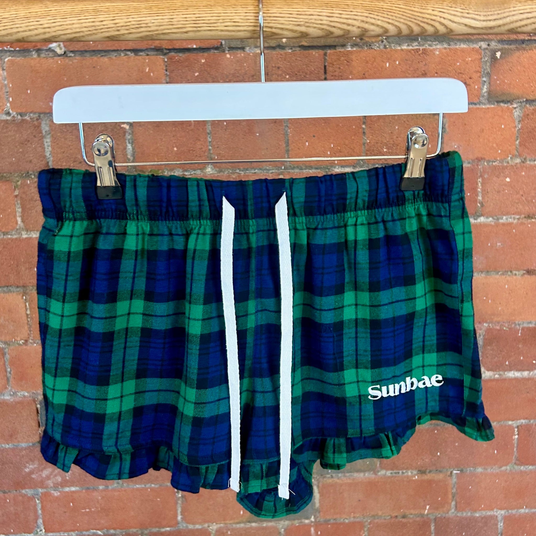 Green plaid shorts with 'Sunbae' branding on a hanger against a brick wall.