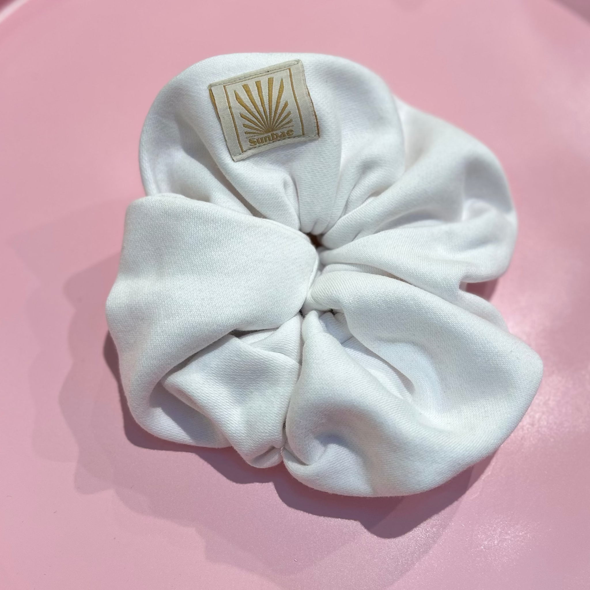 White SUNBAE scrunchie with a SUNBAE label on a pink background.