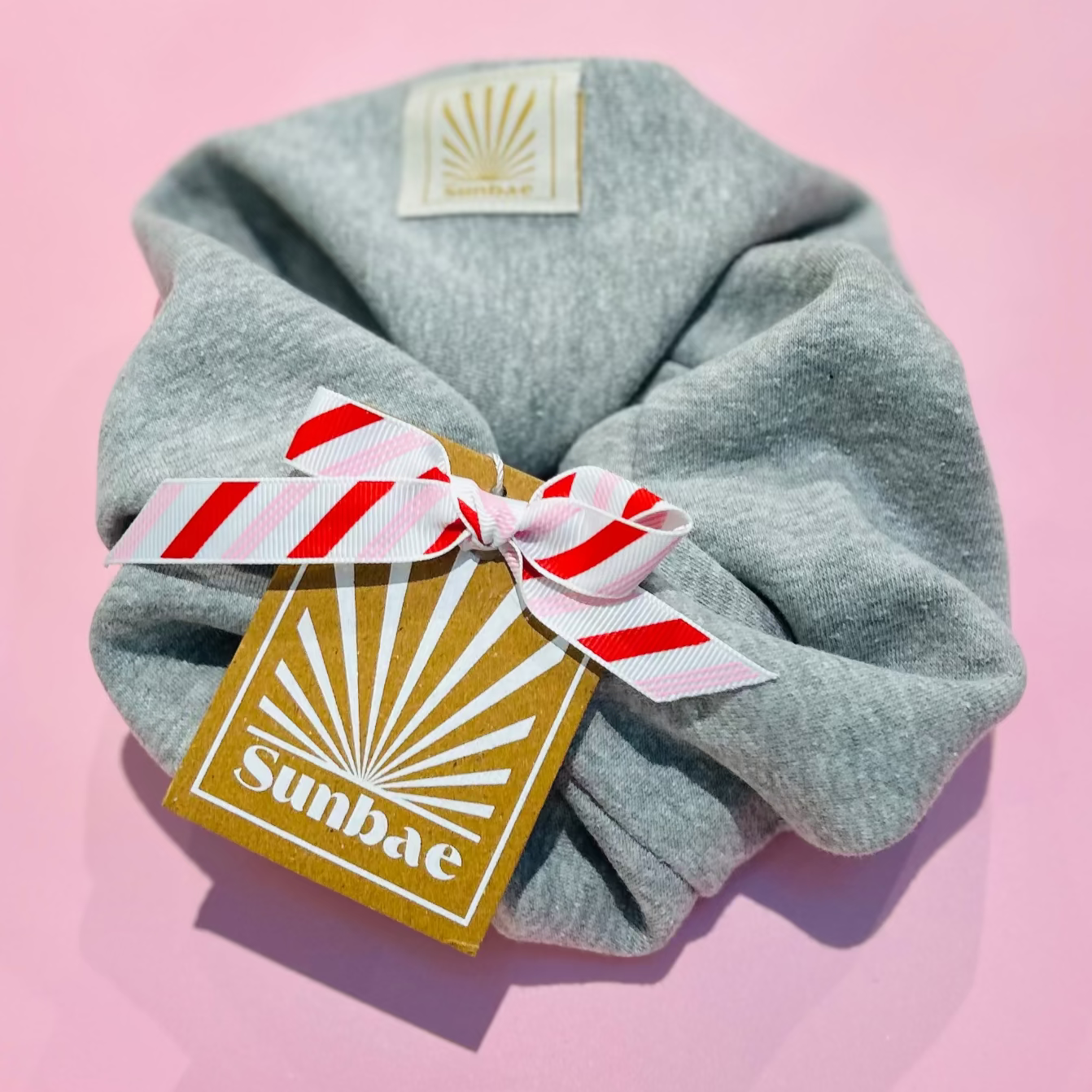 Grey Marl SUNBAE scrunchie with a red and white striped bow SUNBAE tag on a pink surface.