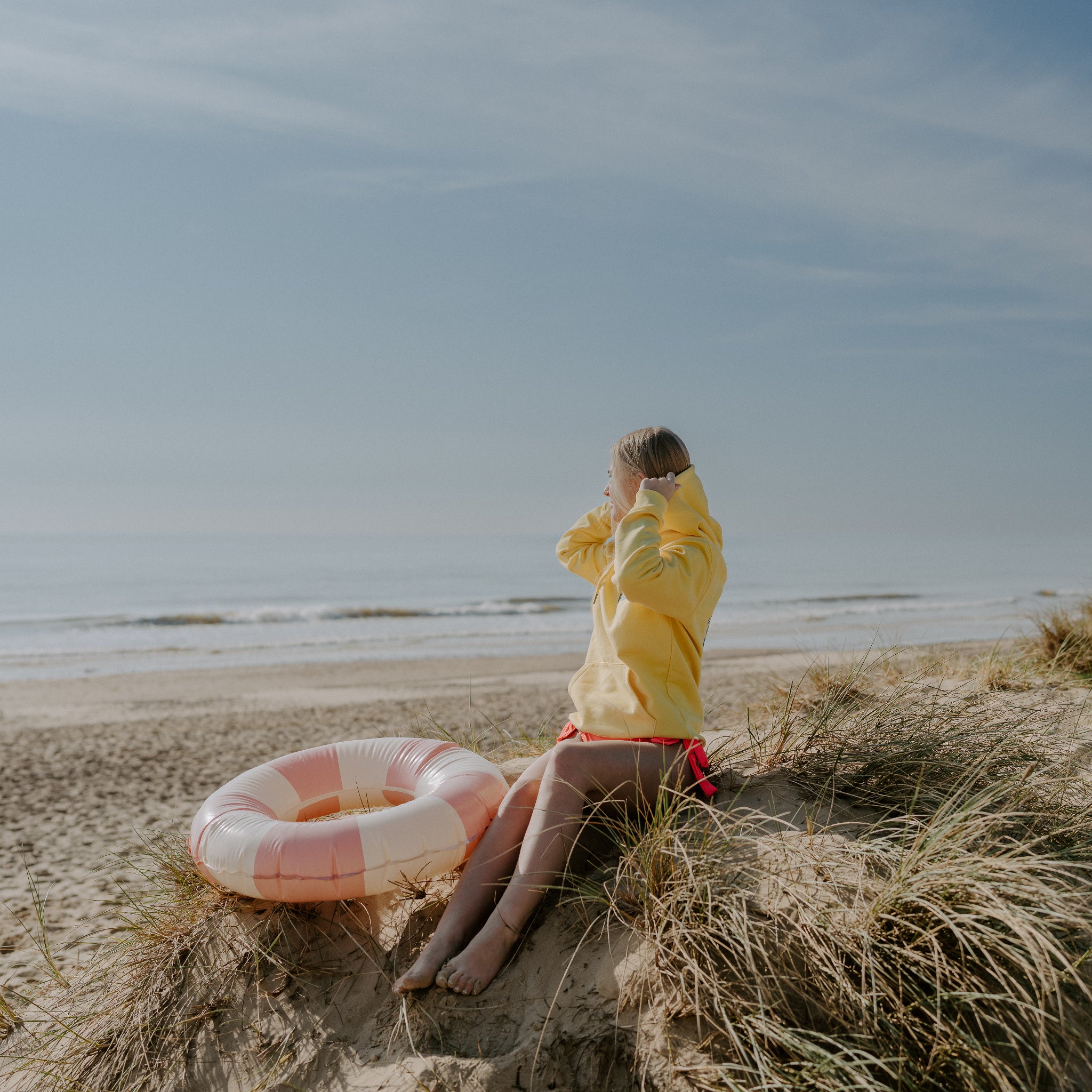 Person in yellow Sunbae hoodie sitting on a beach with a life ring nearby