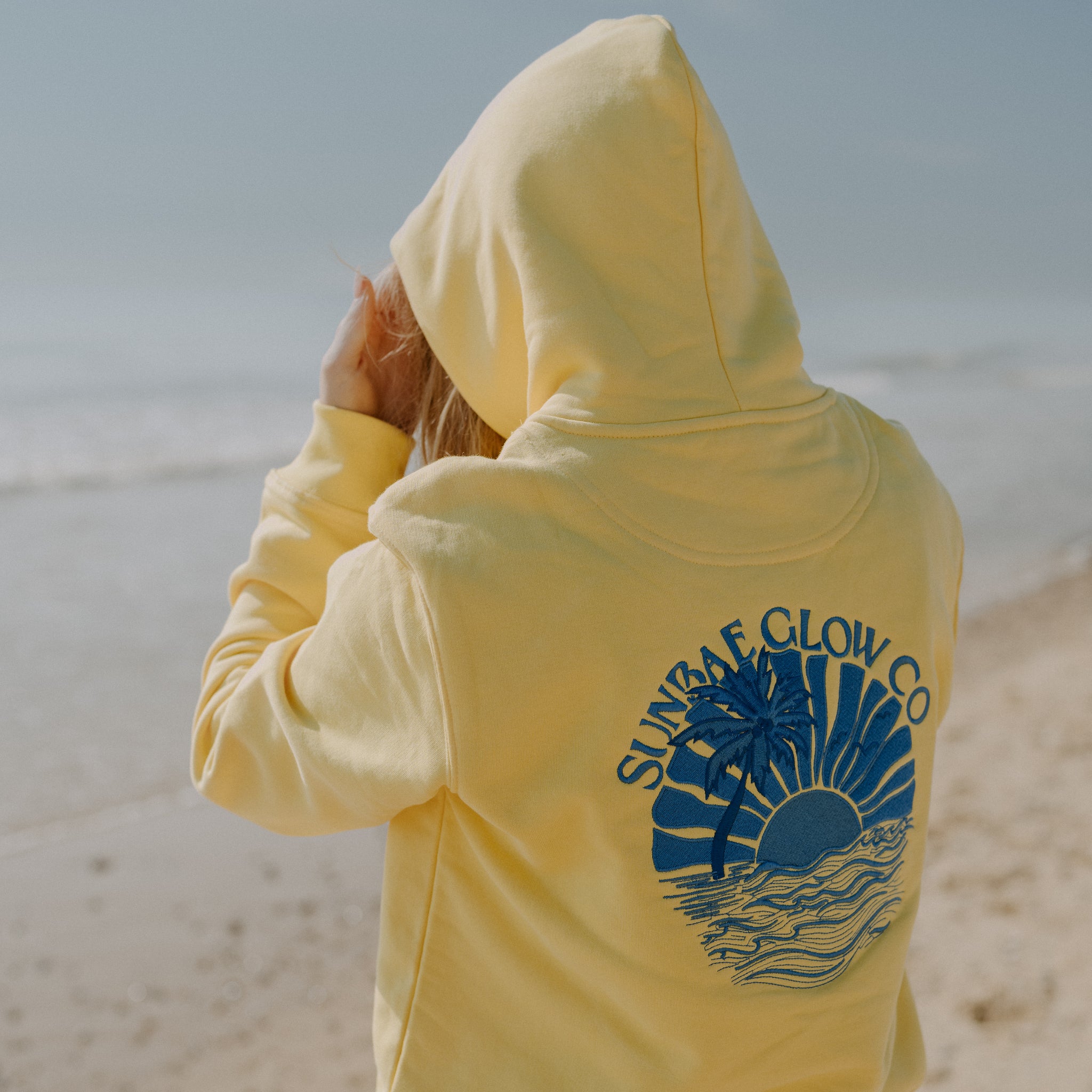 Person wearing a yellow hoodie with 'Sunbae Glow Co' logo on a beach
