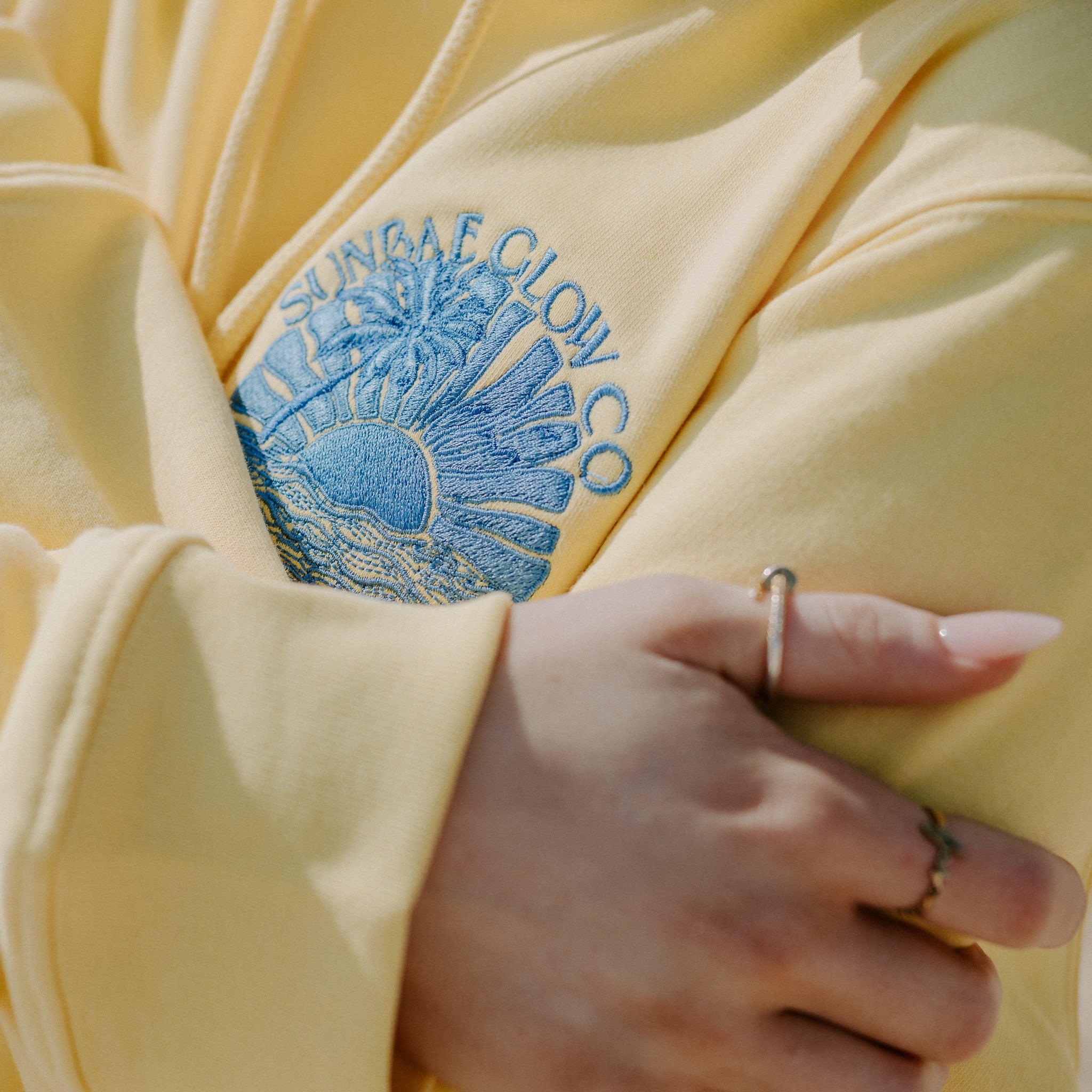 Yellow Sunbae Hoodie with blue embroidered logo and text, held by a hand.