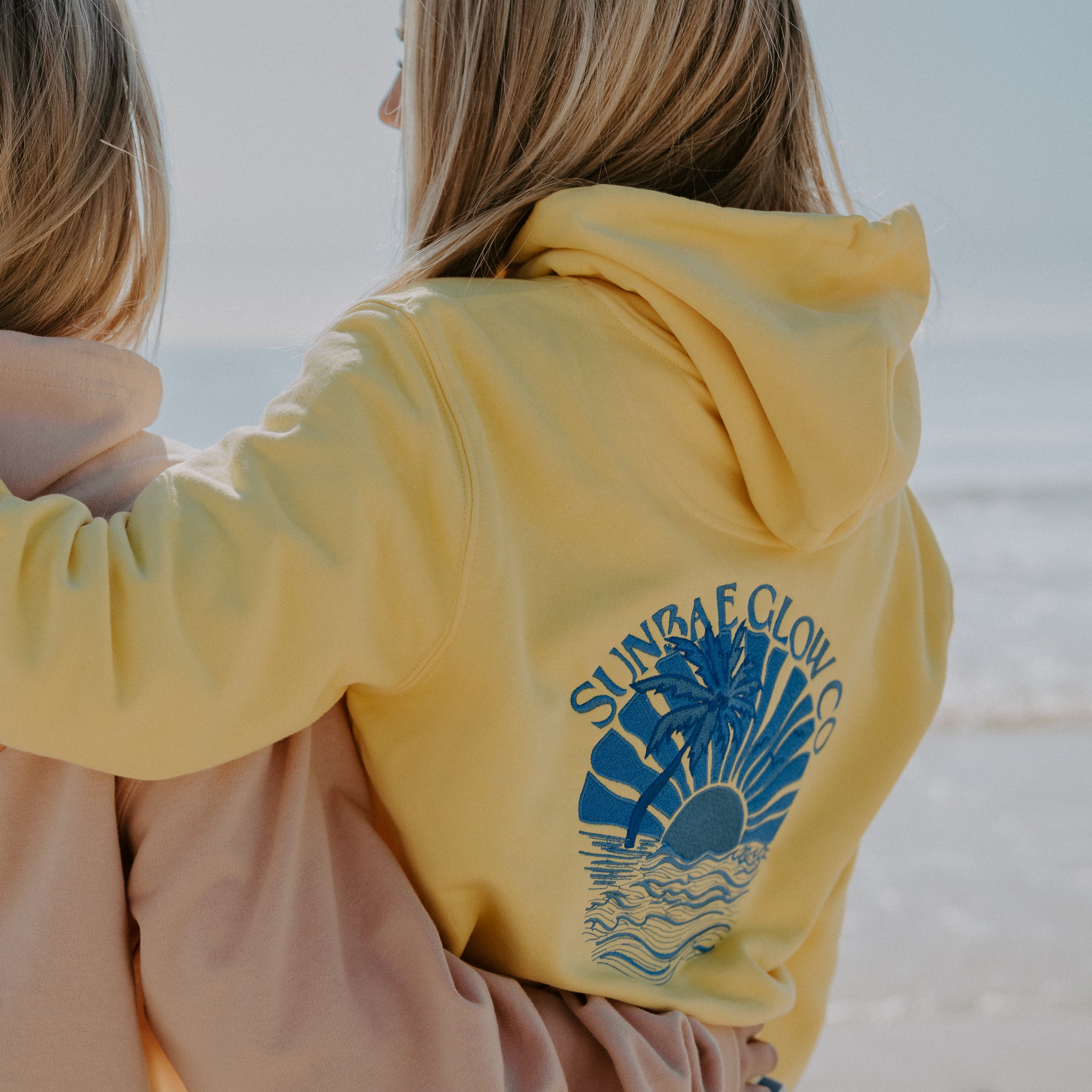Two individuals wearing oversized yellow hoodies with a 'SUNBAE Tidal Glow' graphic on the back, at a beach setting.