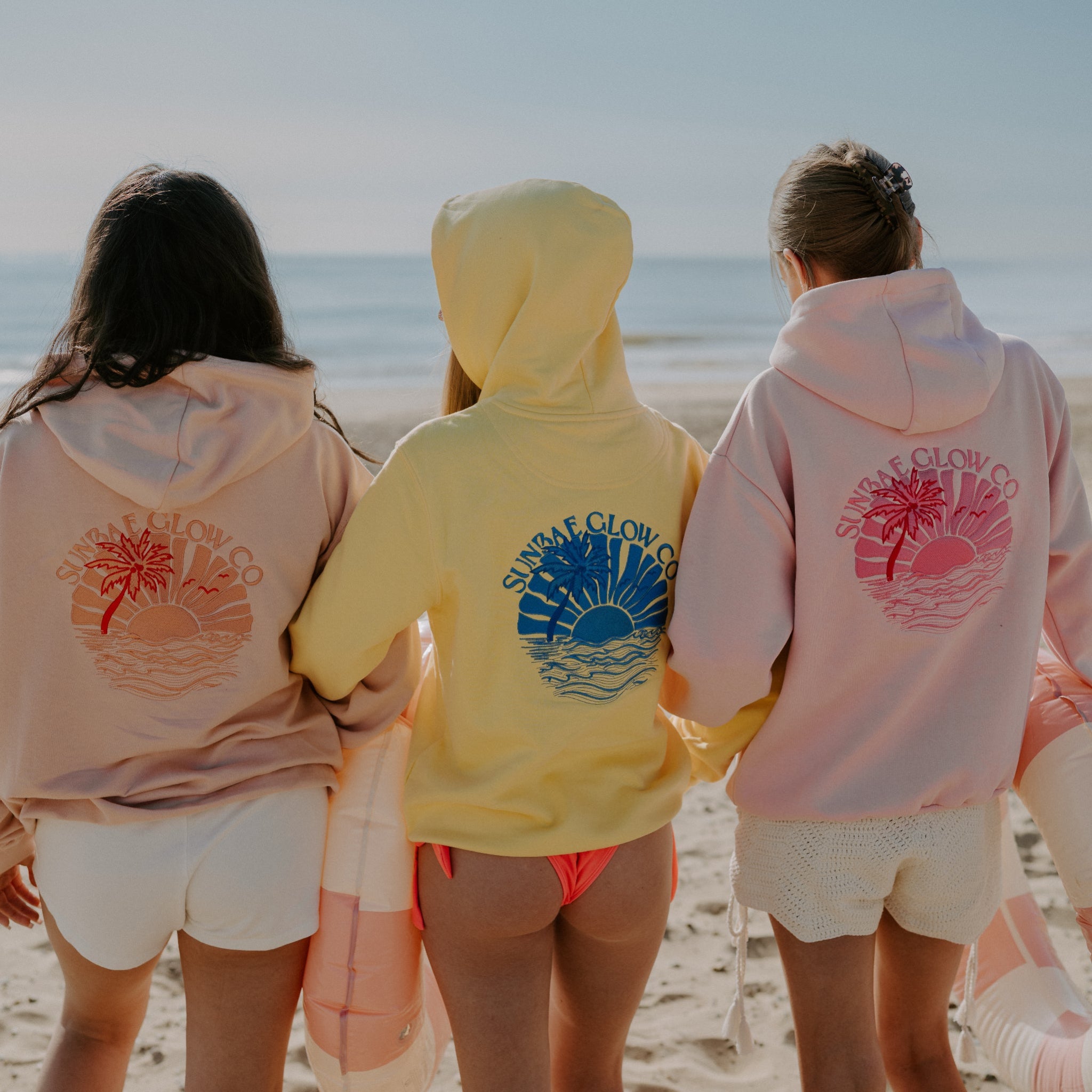 Three women standing on a beach, wearing SUNBAE hoodies in Coral, Yellow and Pink tones, facing away from the camera.