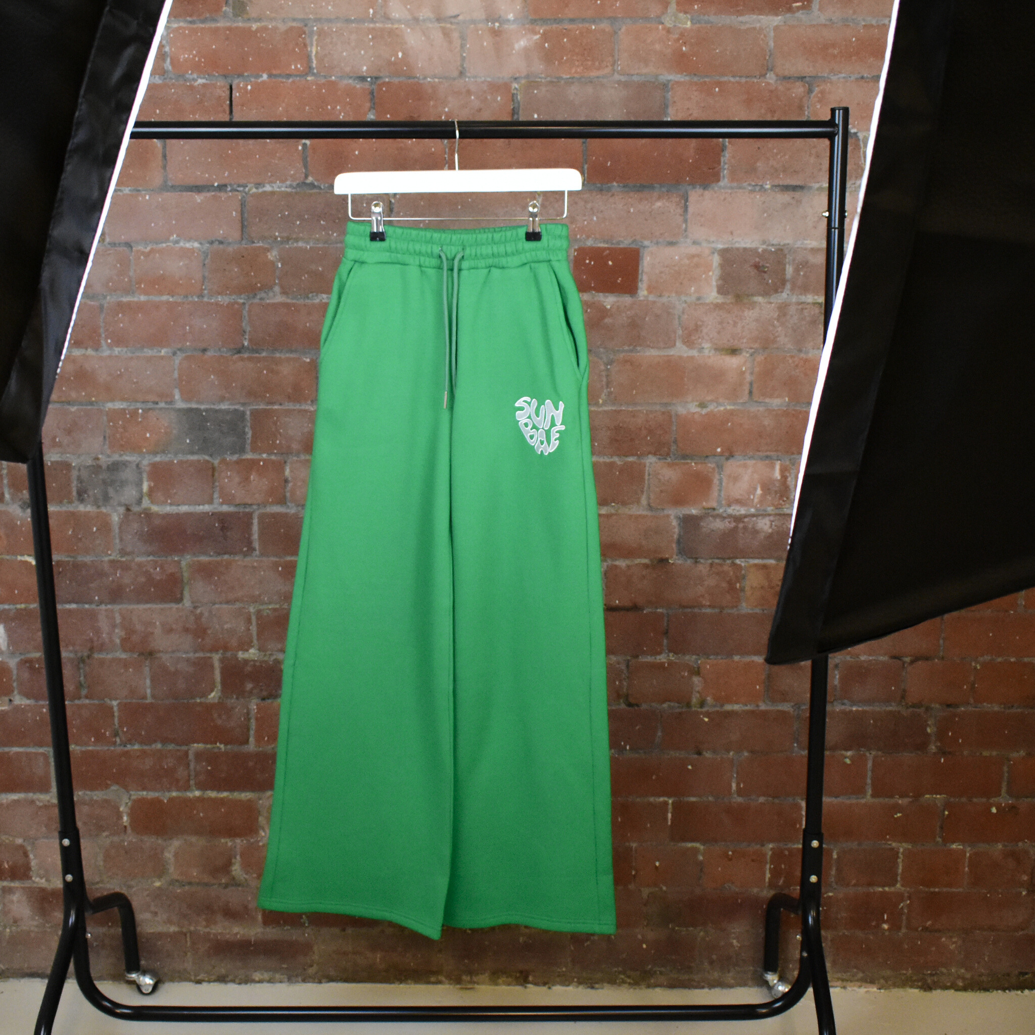 Green SUNBAE Sweat Pants on a hanger against a brick wall