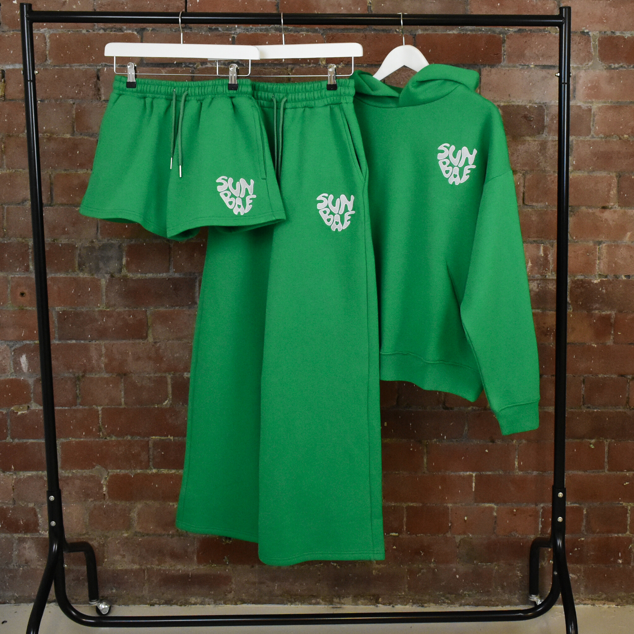 Green SUNBAE hoodie, sweatpants, and shorts set with logo on a rack against a brick wall