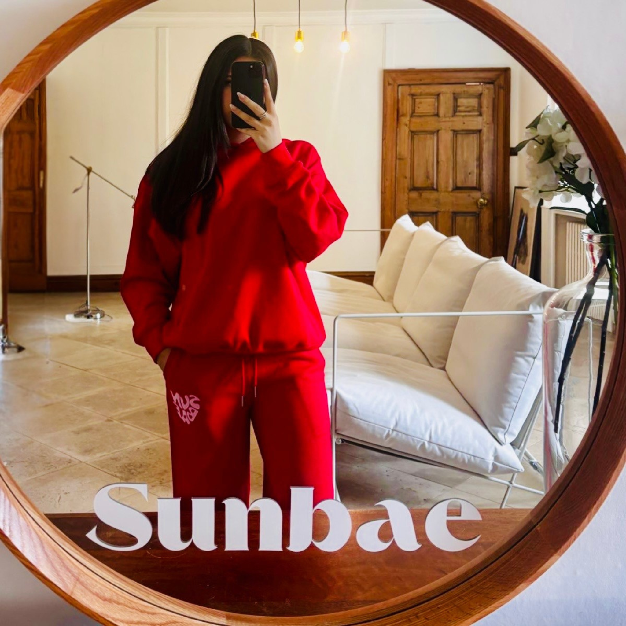 Woman wearing a red Hoodie and Sweat Pant with SUNBAE branding in a living room.