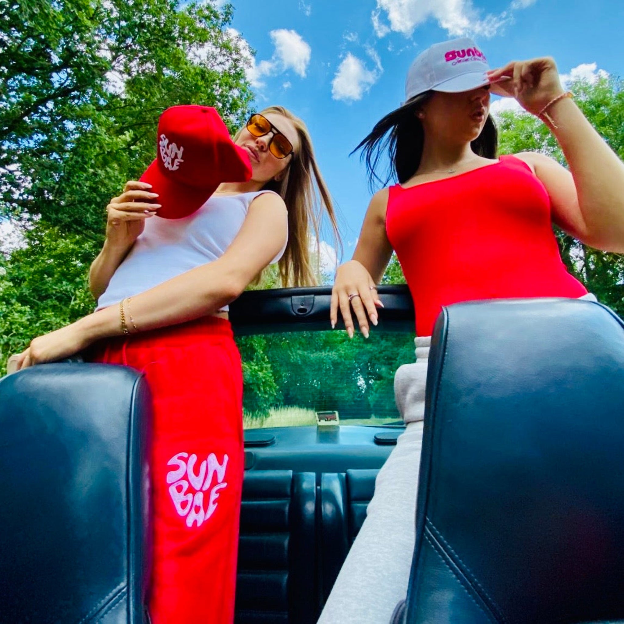 Two women in a car wearing red SUNBAE caps and Sweat Pants with a blue sky and trees in the background.