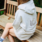Person wearing a light-colored hoodie with a nature design, sitting on a wooden bench outdoors.