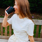 Woman drinking from a black coffee cup while sitting on a wooden bench outdoors.