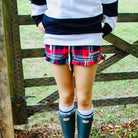 Person wearing a plaid skirt, white sweater, and green wellington boots standing in front of a wooden fence.