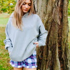 Woman in a gray oversized sweater and plaid skirt standing next to a tree in an outdoor setting