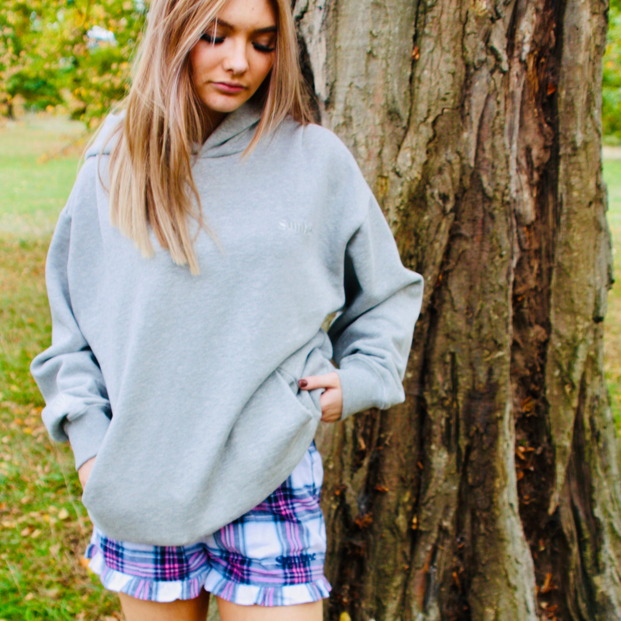 Woman in a gray oversized sweater and plaid skirt standing next to a tree in an outdoor setting