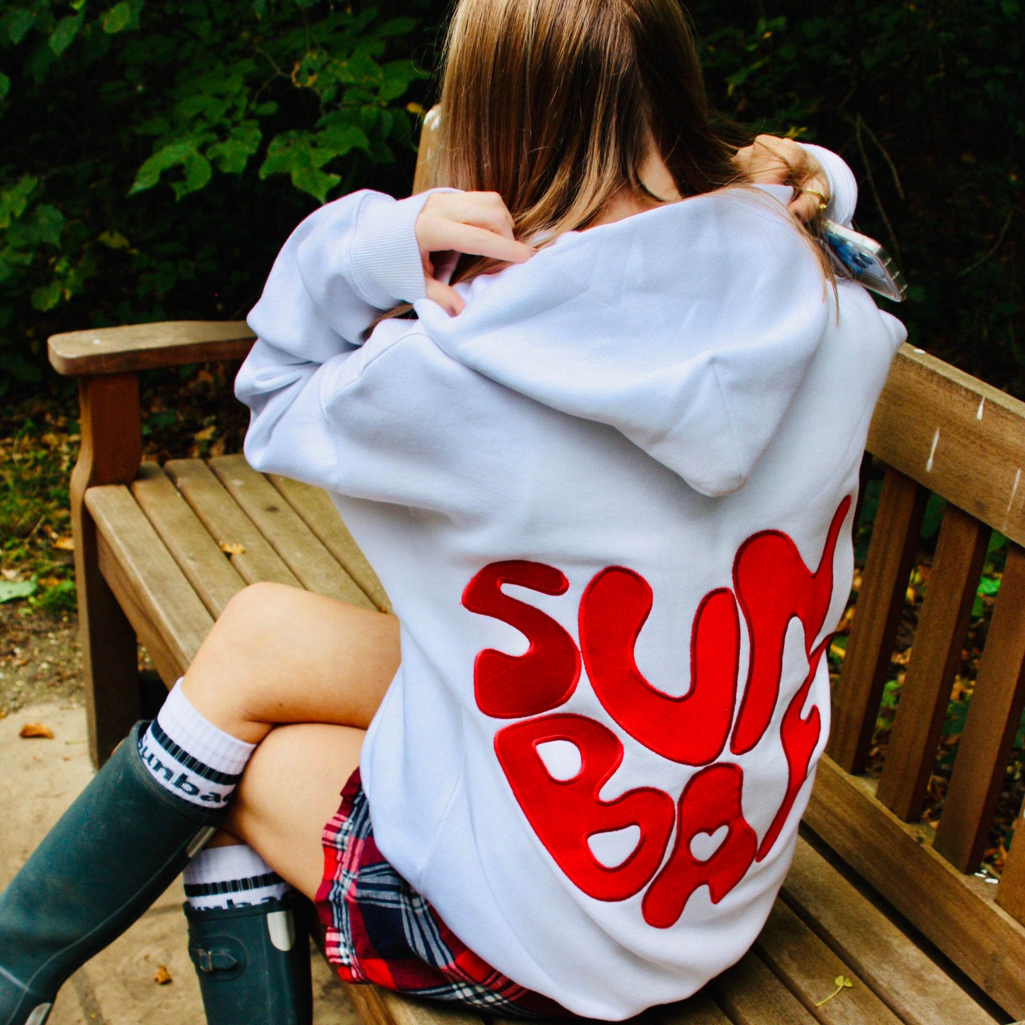 Person wearing a hoodie with 'Sun Bum' text sitting on a wooden bench outdoors.