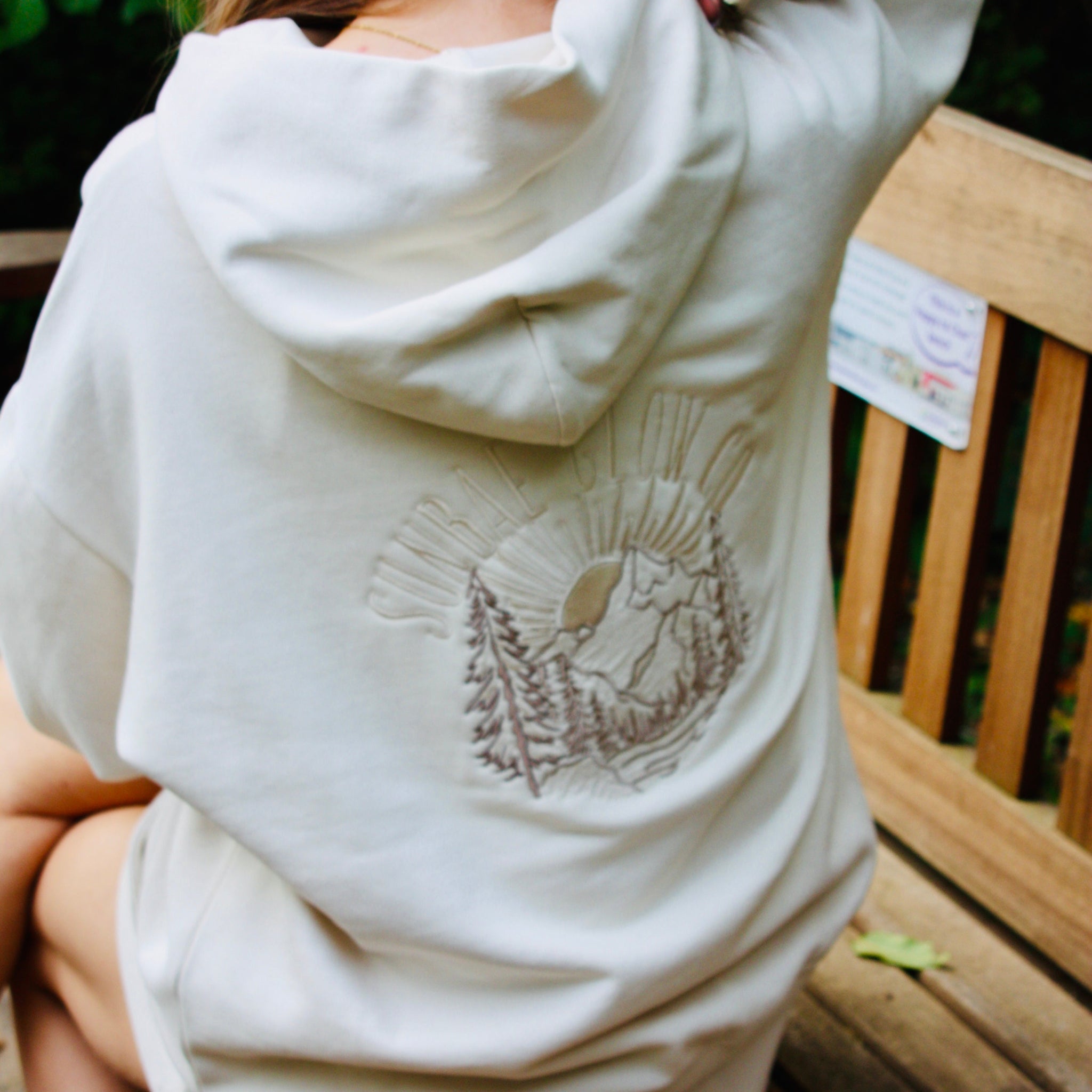 Person wearing a white hoodie with a graphic design sitting on a wooden bench outdoors.