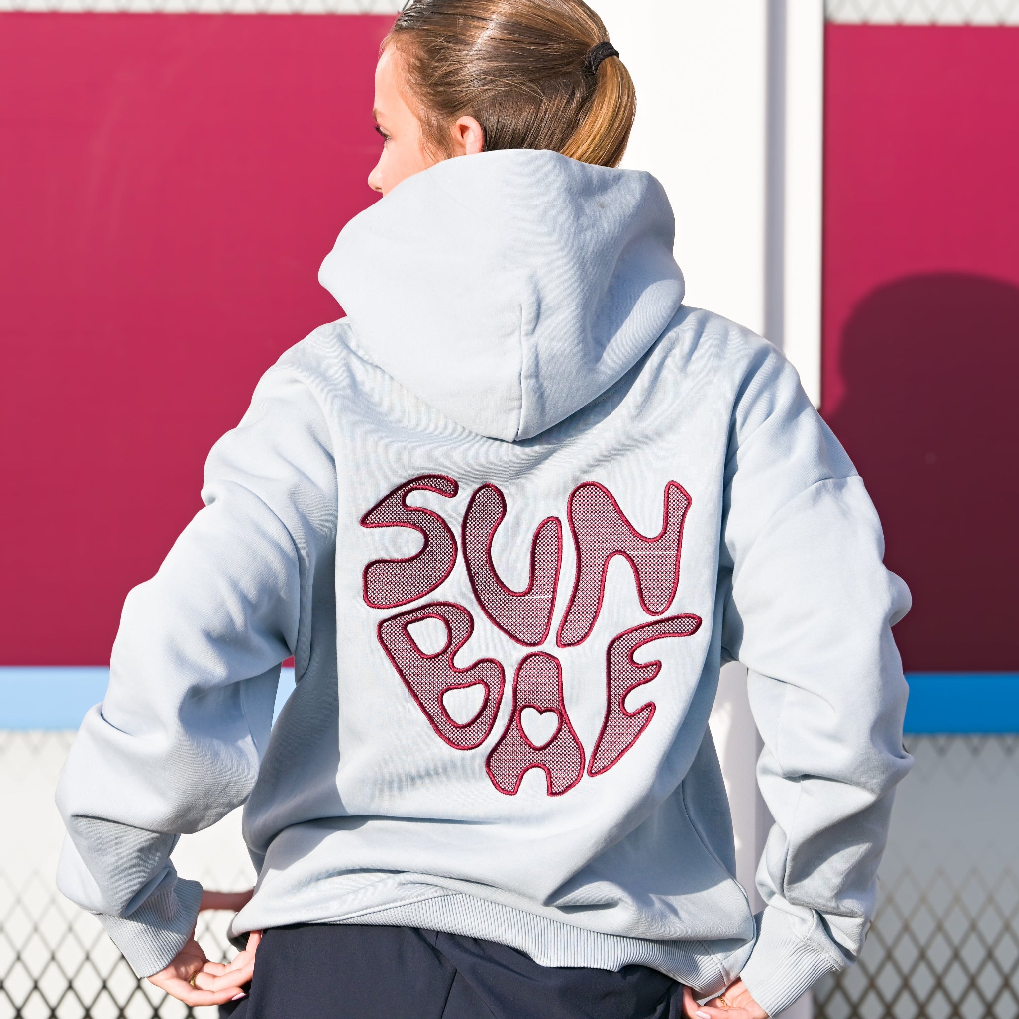 Yvonne Main wearing a Sunbae hoodie wiith text on the back, standing in front of a red wall.