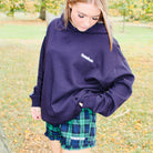 Person wearing a purple sweatshirt and plaid shorts standing in a park with trees and grass in the background.