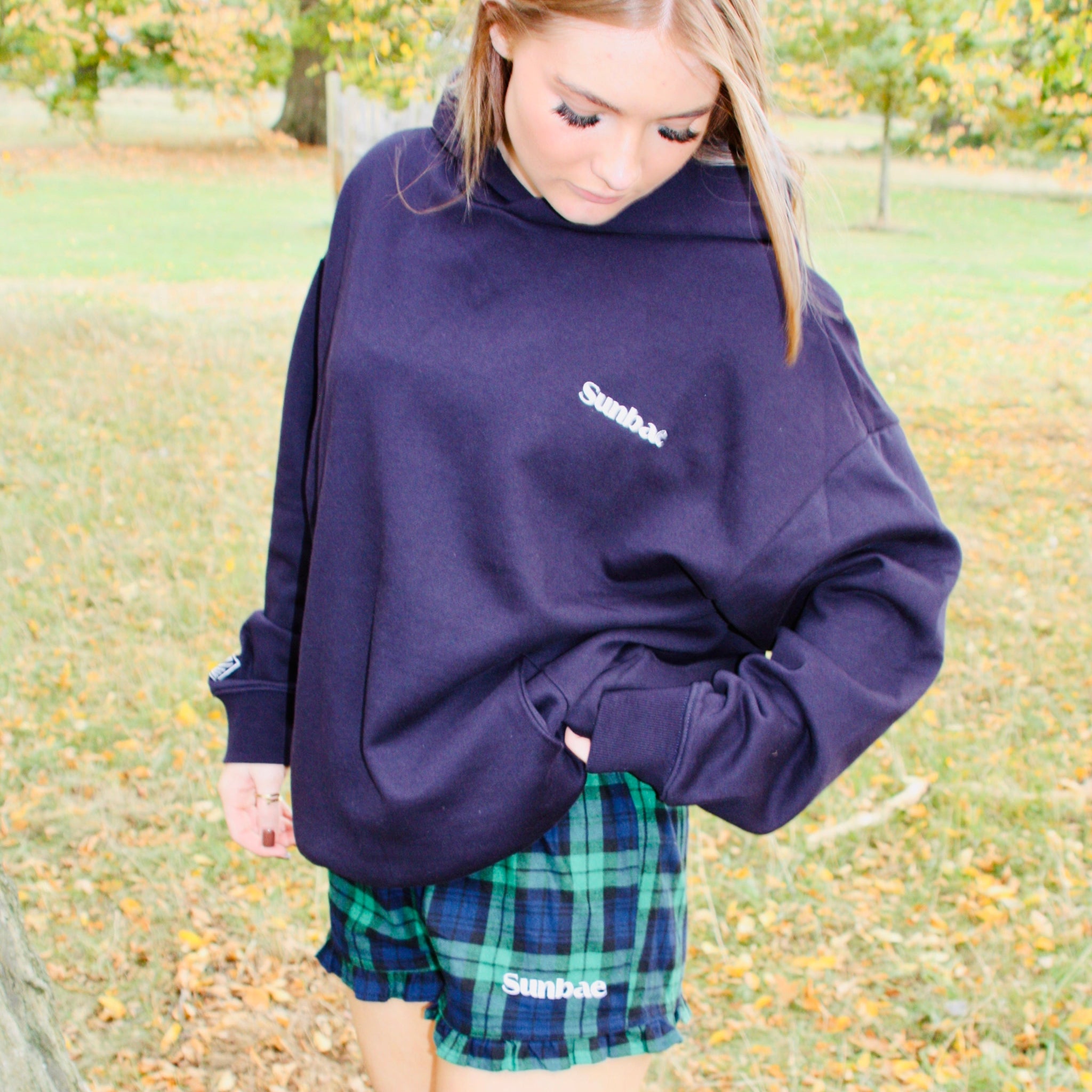 Person wearing a purple sweatshirt and plaid shorts standing in a park with trees and grass in the background.