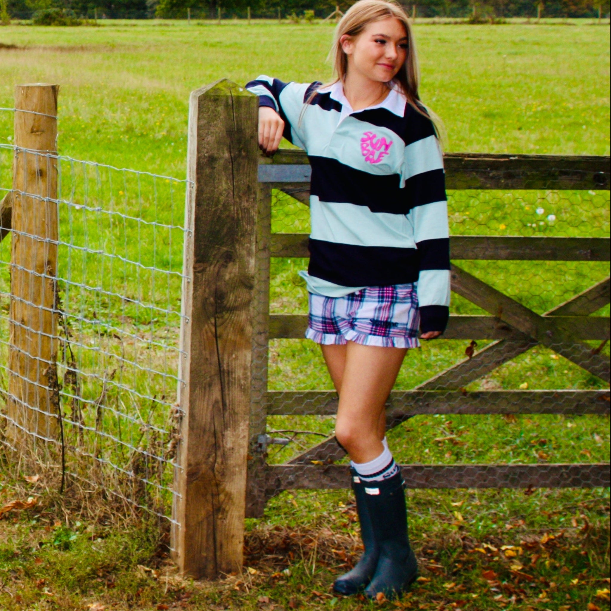 Person standing by a wooden gate in a grassy field with trees in the background