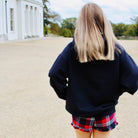 Person wearing a black sweater, plaid skirt, and rain boots standing on a gravel path with trees and a building in the background.