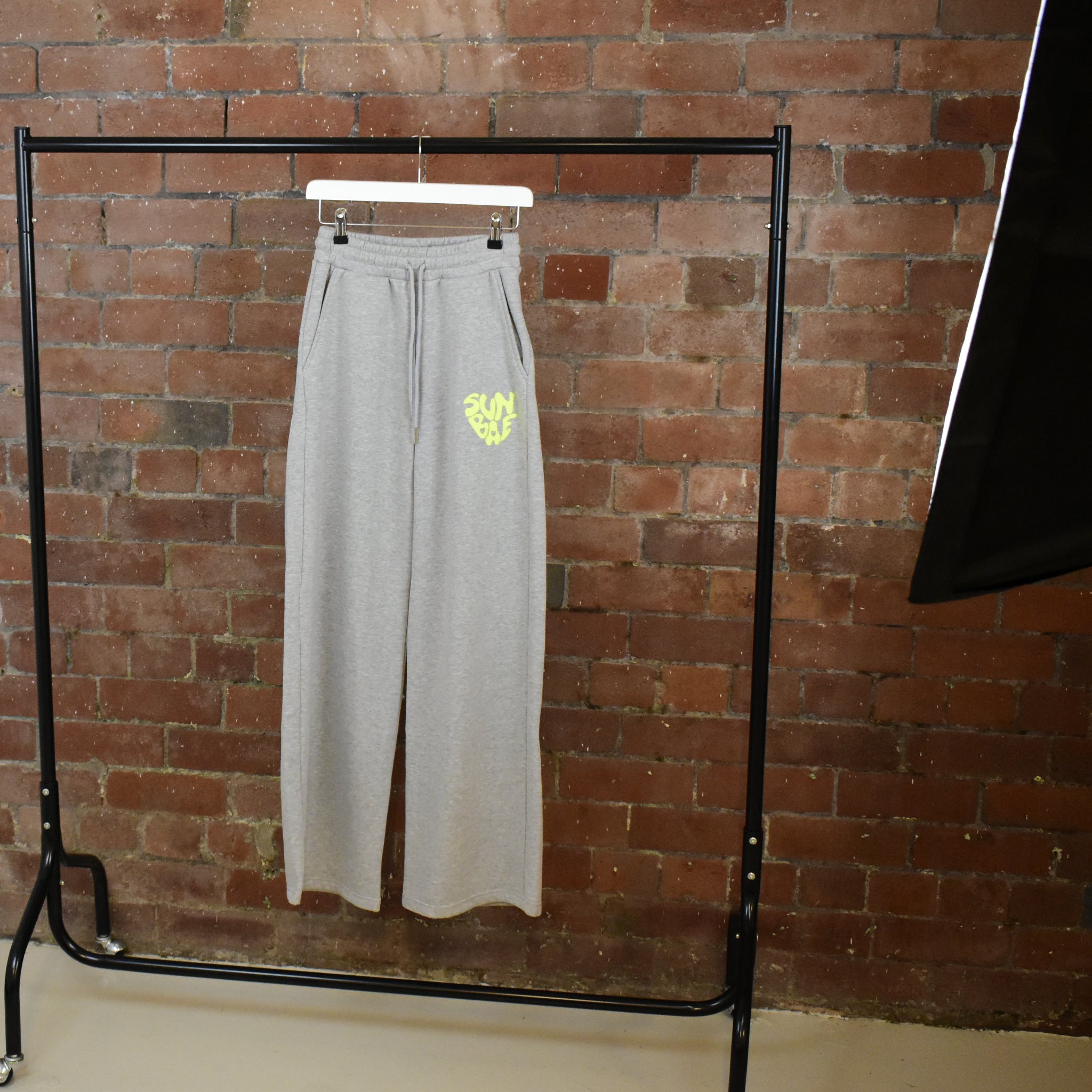 Grey SUNBAE sweatpants with a yellow logo on a hanger against a brick wall.